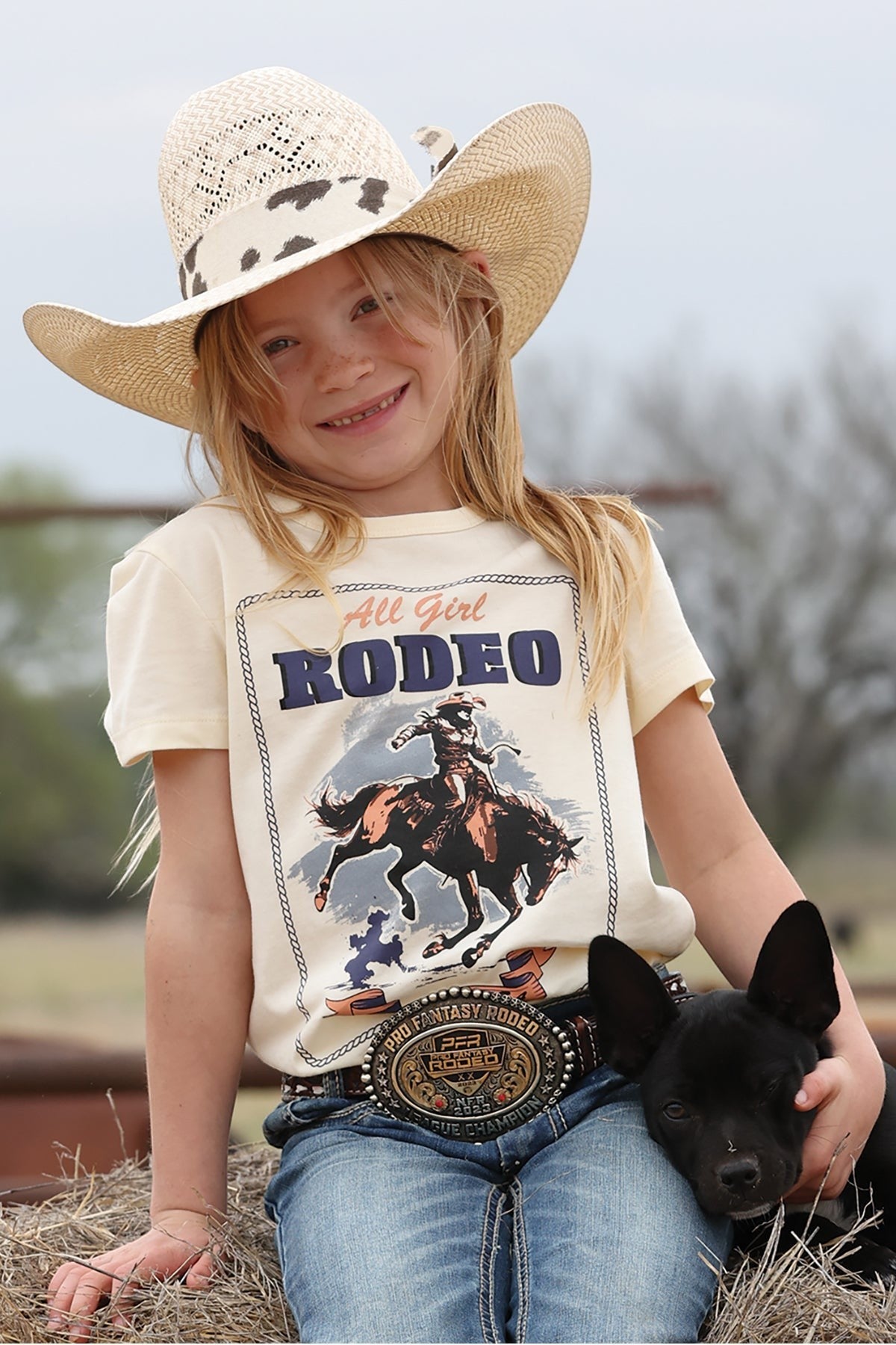 Young girl wearing a 'All Girl Rodeo' t-shirt with a cowboy graphic, holding a black dog, and wearing a cowboy hat.