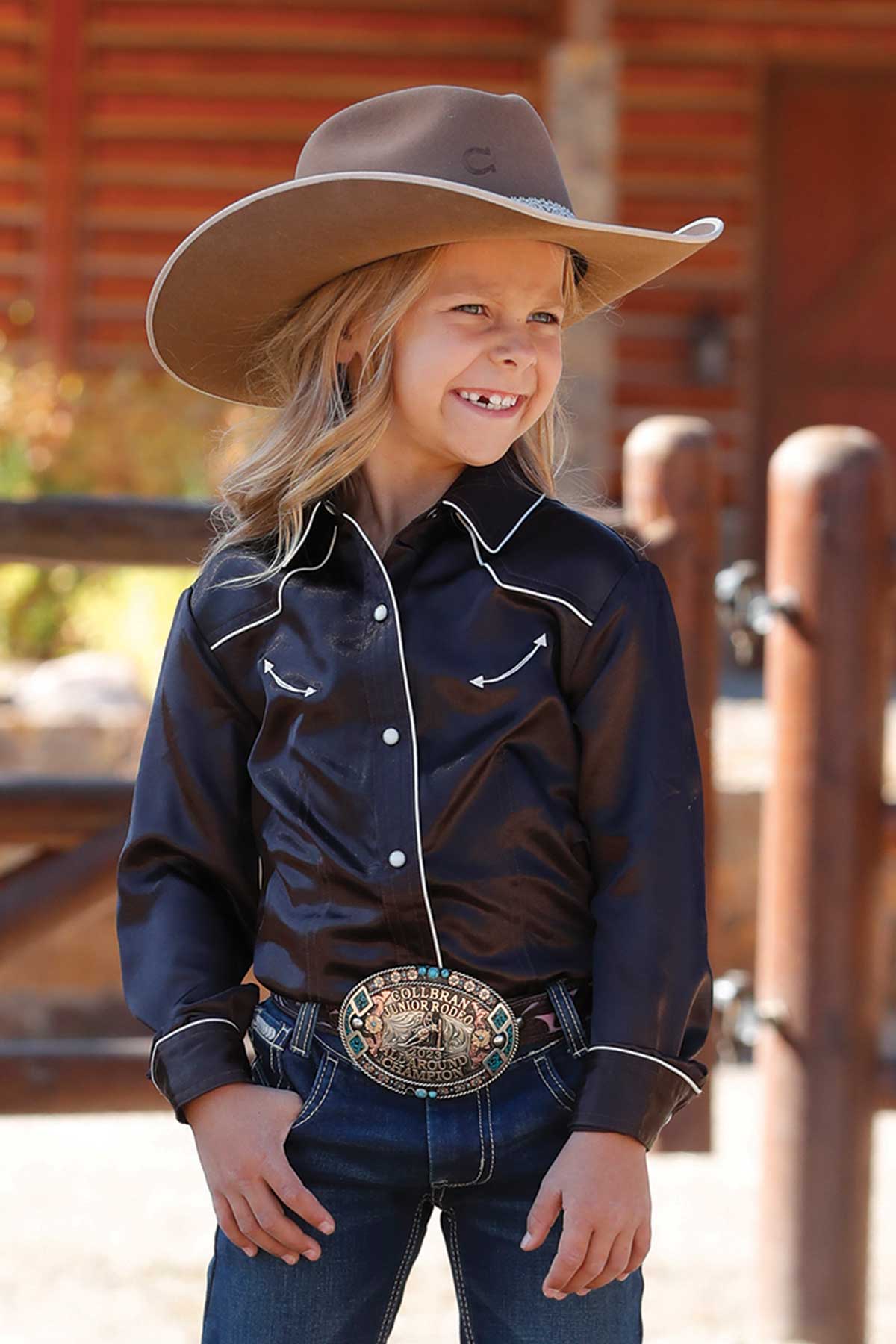 Young girl wearing a dark long sleeve snap-front shirt with white piping details, dark jeans, and a belt with an ornate buckle. She has on a cowboy hat and is smiling while standing outside near a wooden fence with a log cabin in the background