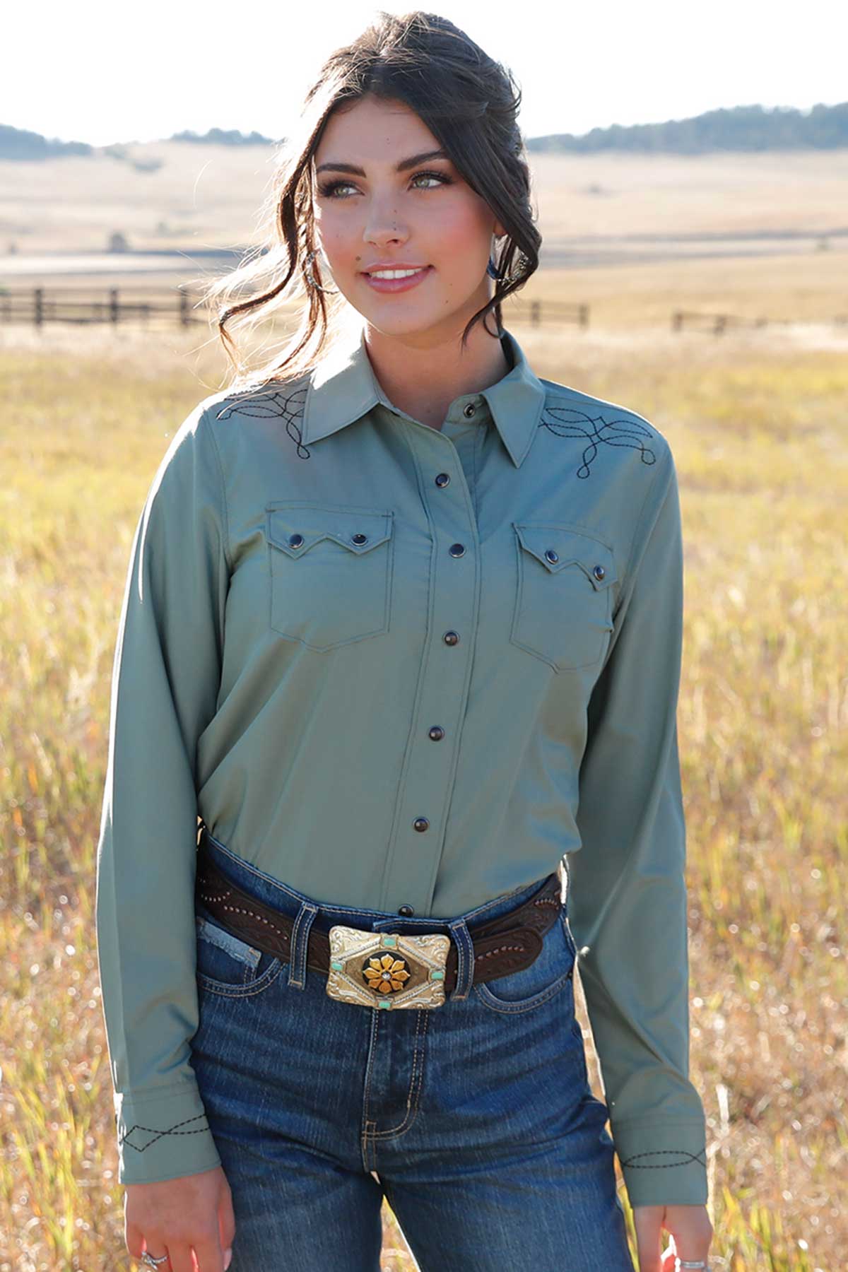 Woman wearing a sage green long-sleeve western shirt with black embroidery, paired with blue jeans and a decorative belt buckle, standing in a grassy field