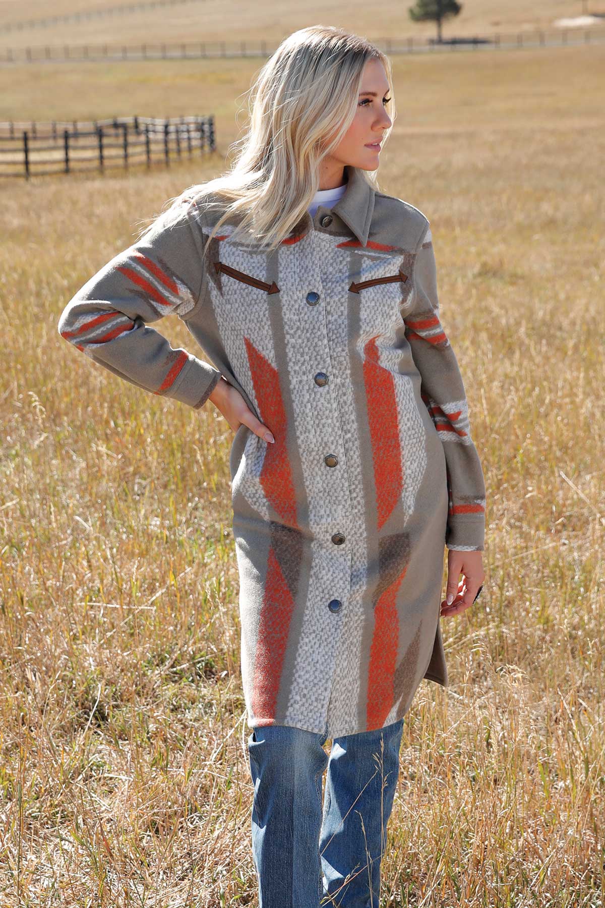 Woman standing in a field wearing a long button-up coat with a Southwestern-inspired pattern in shades of gray, rust, and cream. The coat has front pockets and a structured collar, paired with blue jeans. The background shows a fence and open pasture