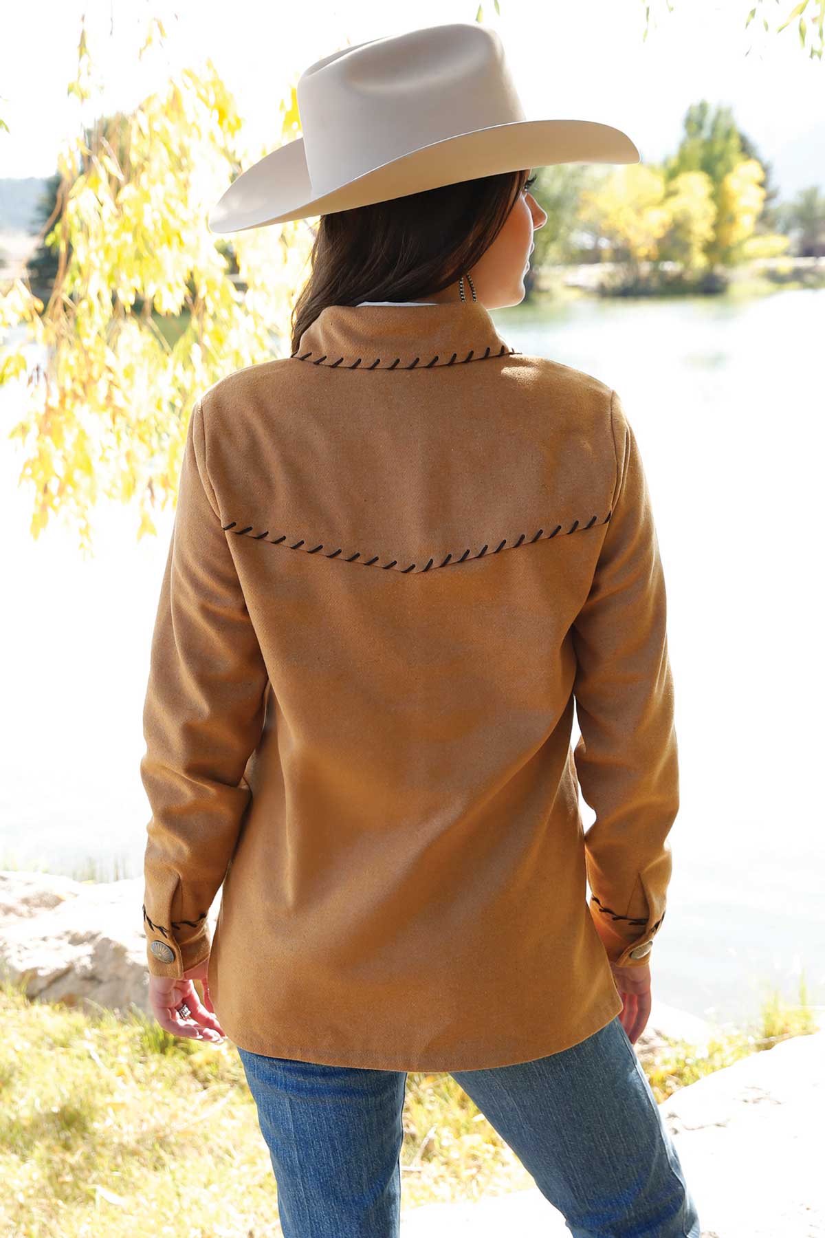Back view of a woman wearing a tan Western-style jacket with black decorative stitching across the shoulders. The outfit is paired with blue jeans and a light-colored cowboy hat, with trees and a lake in the background