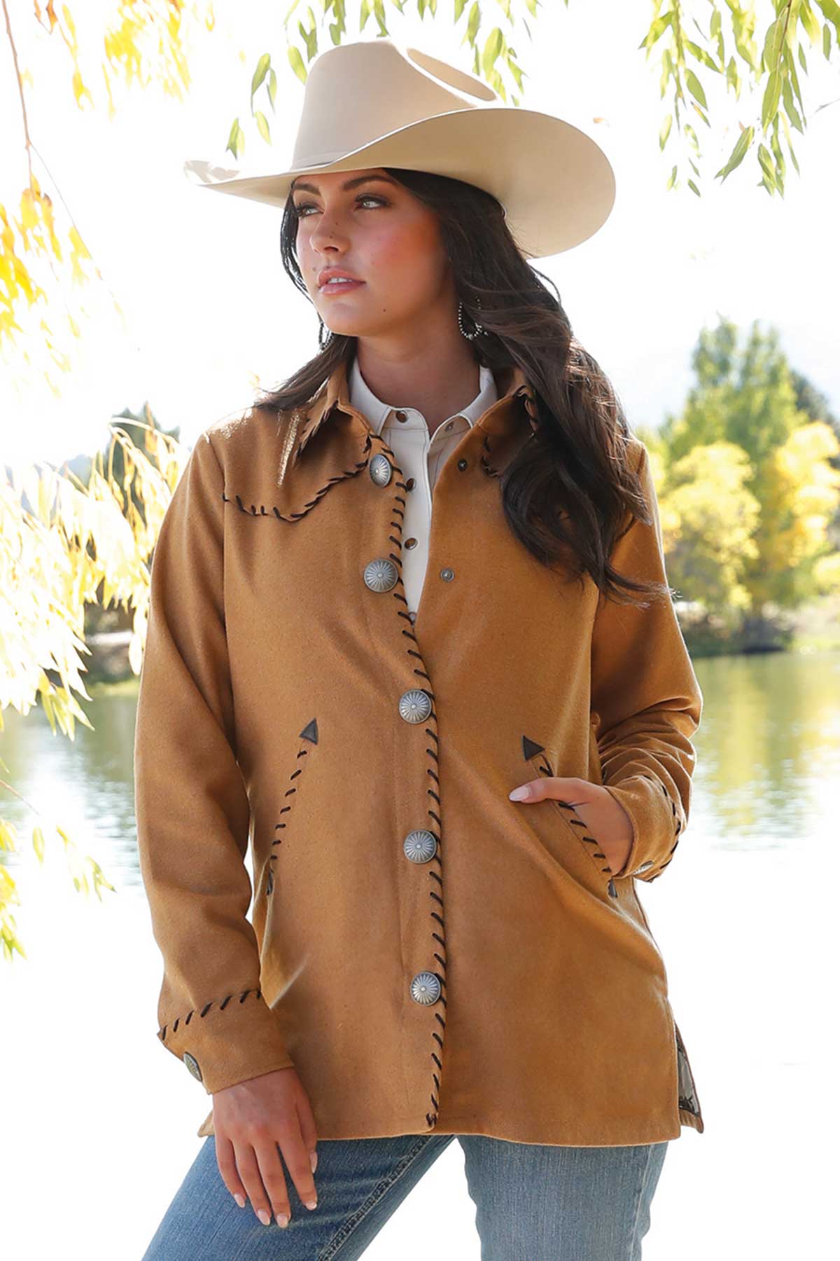 Woman wearing a tan Western-style jacket with decorative black stitching and large silver concho buttons, paired with blue jeans and a white cowboy hat, standing outdoors near a lake with sunlight filtering through tree branches