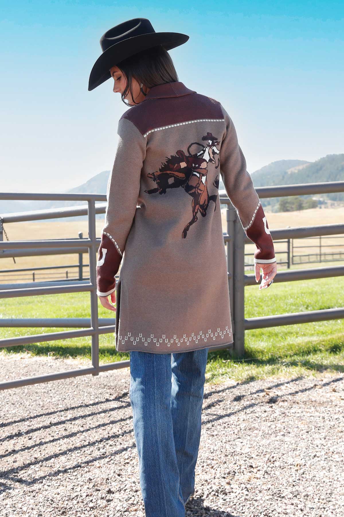 Woman facing away, wearing a long brown and burgundy cardigan with a detailed bucking horse and rider graphic on the back, paired with blue jeans and a black cowboy hat, standing near a fence with fields and hills in the background
