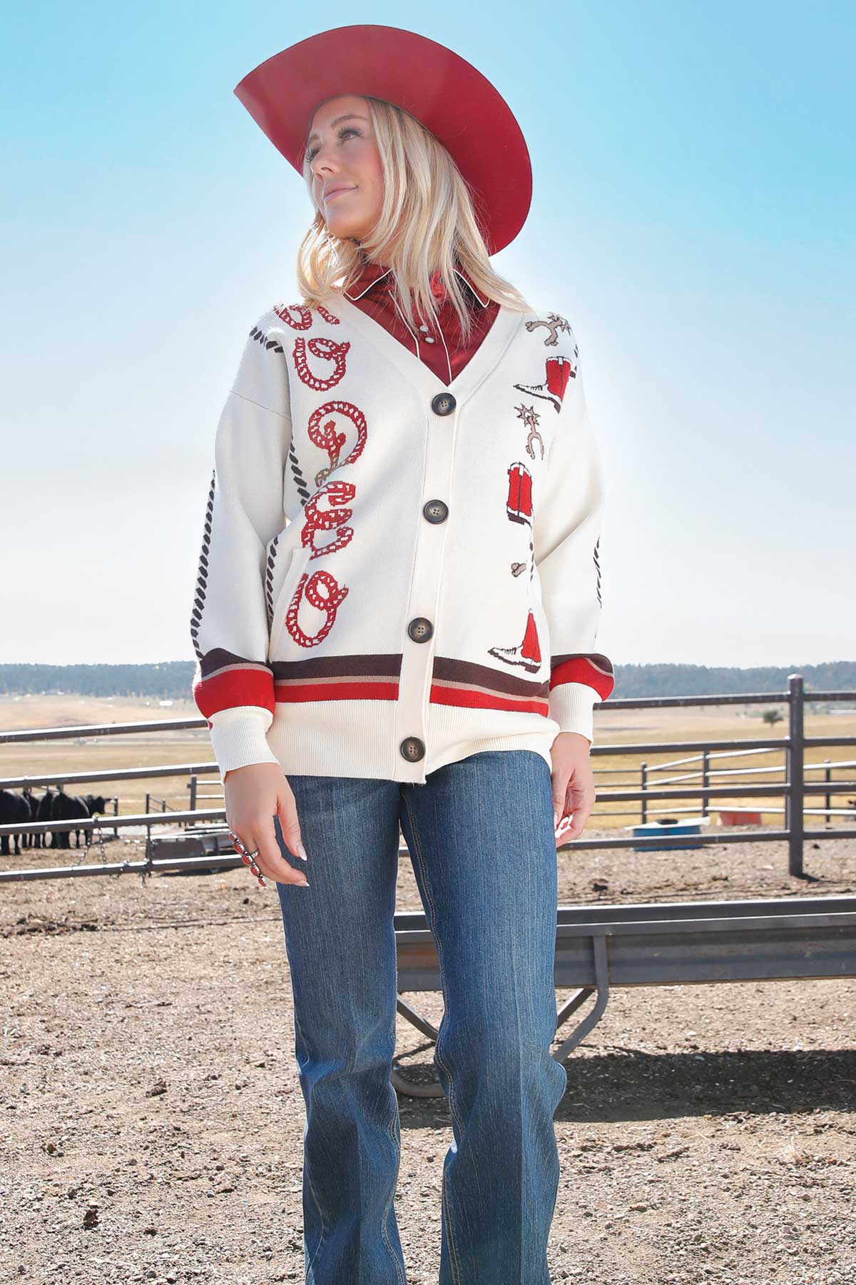 Girl wearing a red cowboy hat, red button-up shirt, and cream cardigan with western embroidery, paired with blue jeans, standing outside in an arena