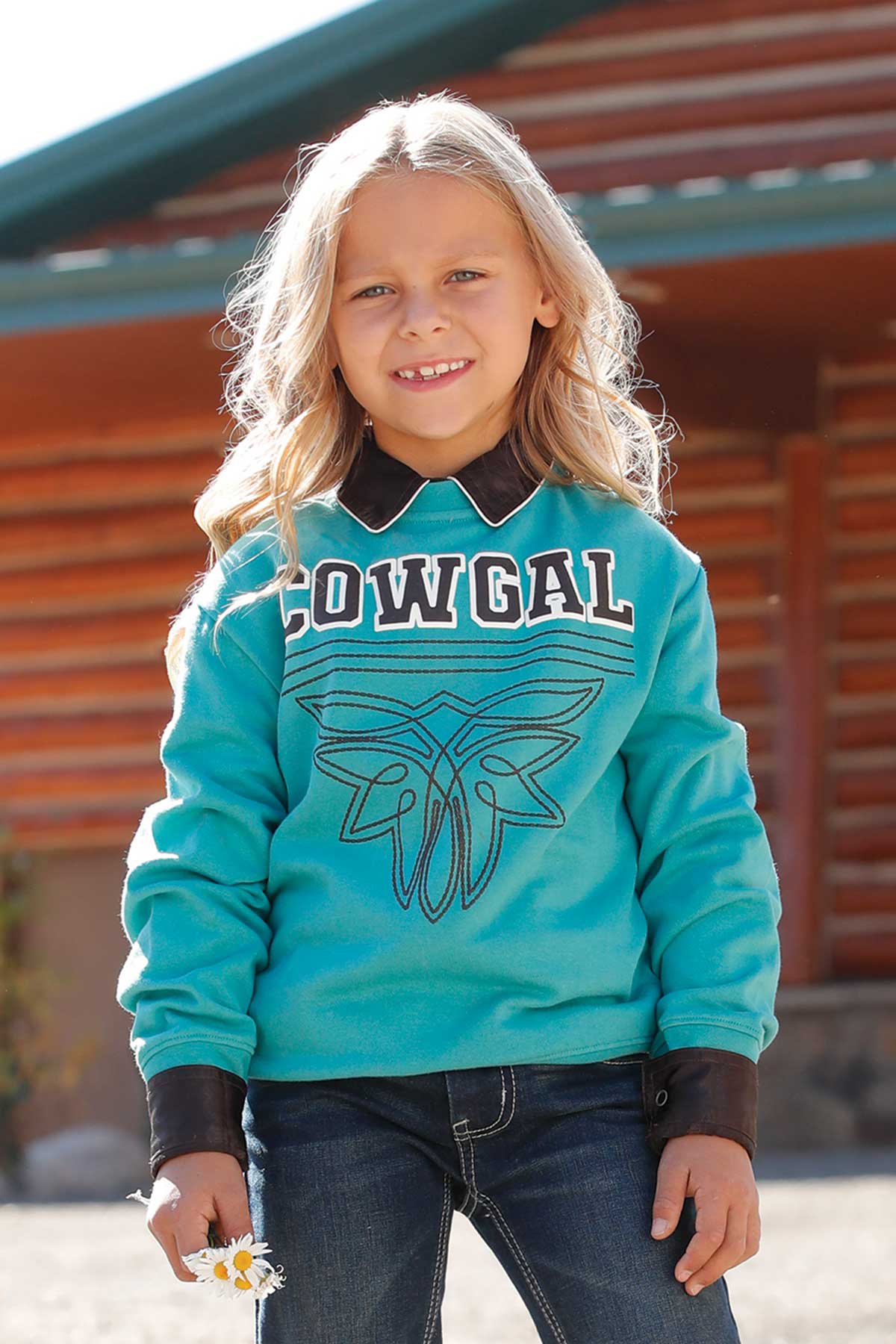 Young girl standing outdoors wearing a turquoise sweatshirt with the word 'COWGAL' in bold lettering and a graphic design beneath it. She has a collared shirt layered underneath, dark jeans, and is holding a small white flower in her hand. A log cabin is visible in the background