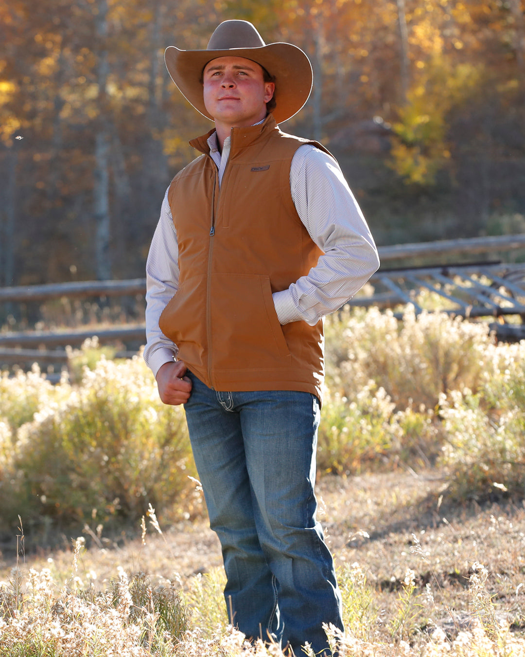 Man wearing a brown vest over a plaid shirt with jeans and a cowboy hat standing outside