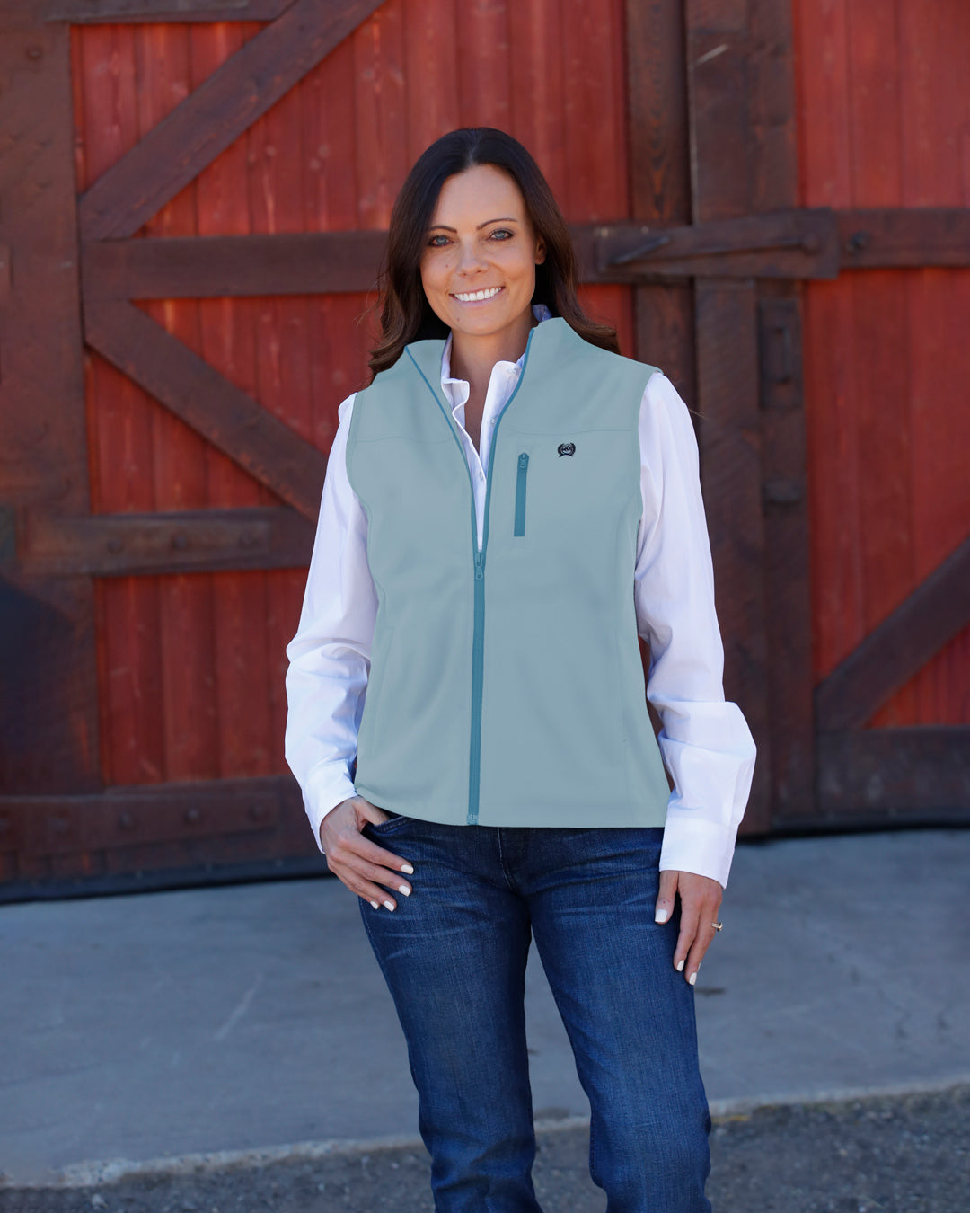 Woman wearing a light blue vest over a white shirt and jeans standing outside in front of a barn
