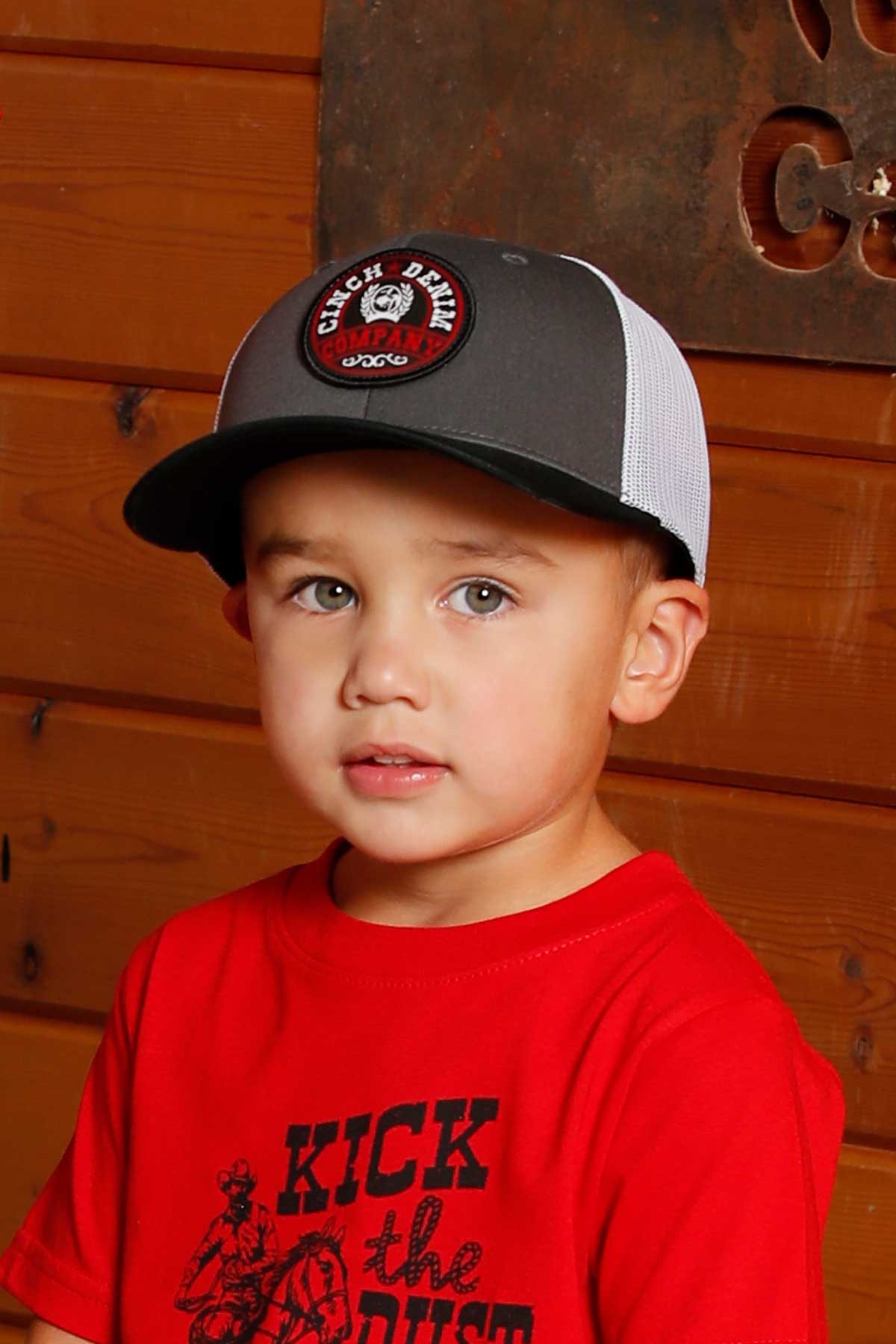 Young boy wearing a red t-shirt and a grey hat, leaning against a wooden wall 