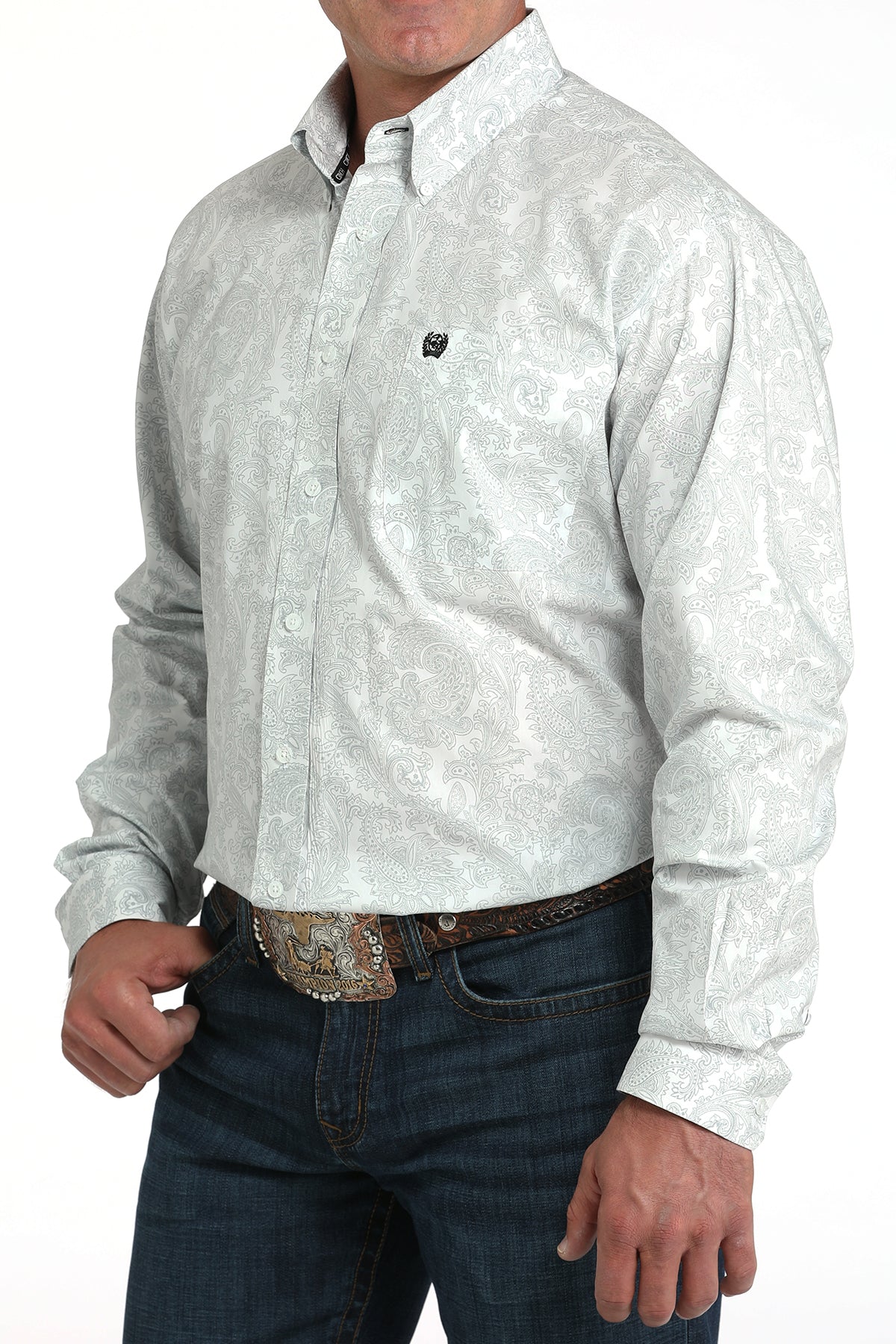 A man wearing a light patterned button-up shirt, dark jeans, and a large belt buckle stands against a white background