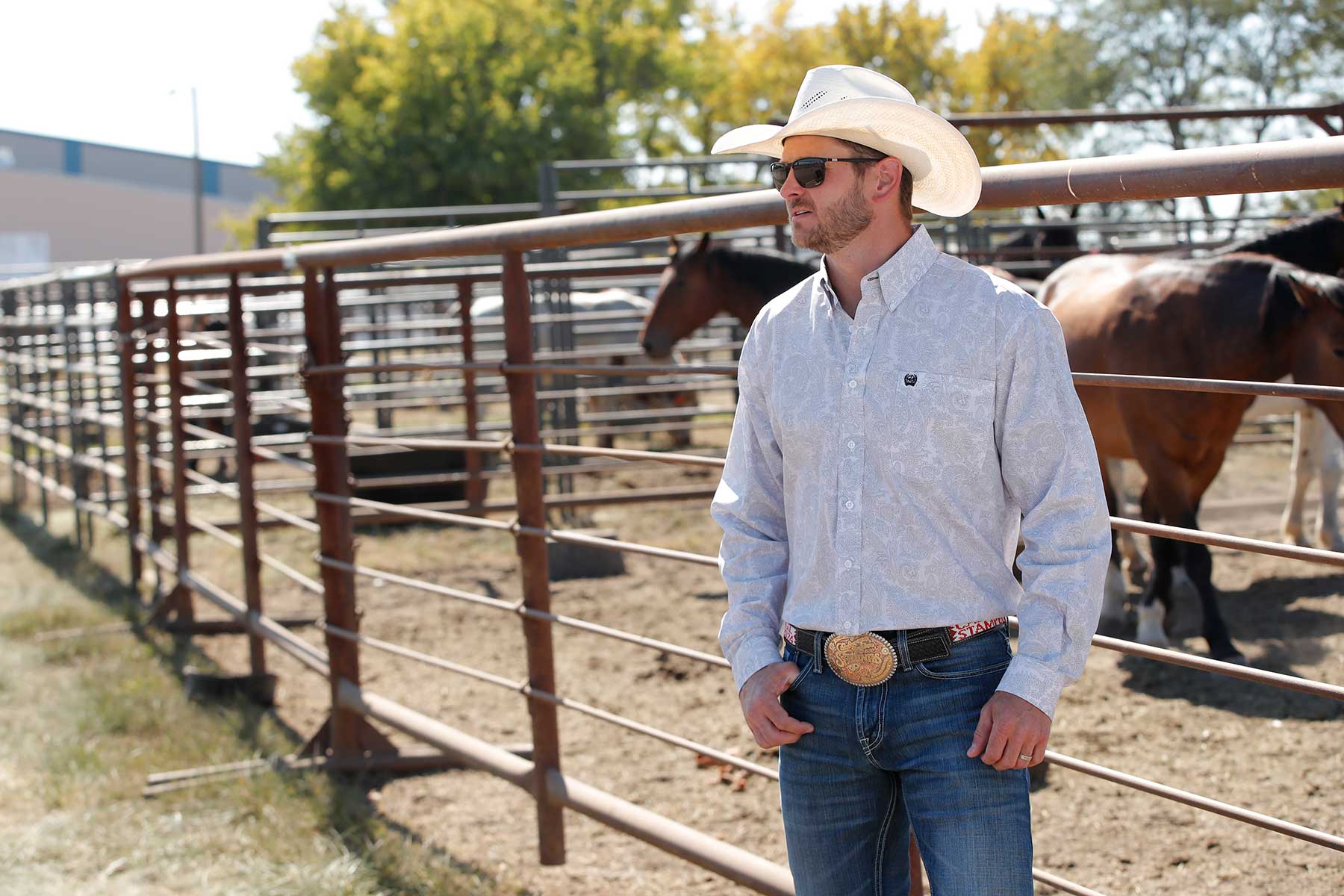 A man wearing a white cowboy hat, patterned long-sleeve shirt, jeans, and a large belt buckle stands by a metal fence with horses in the background