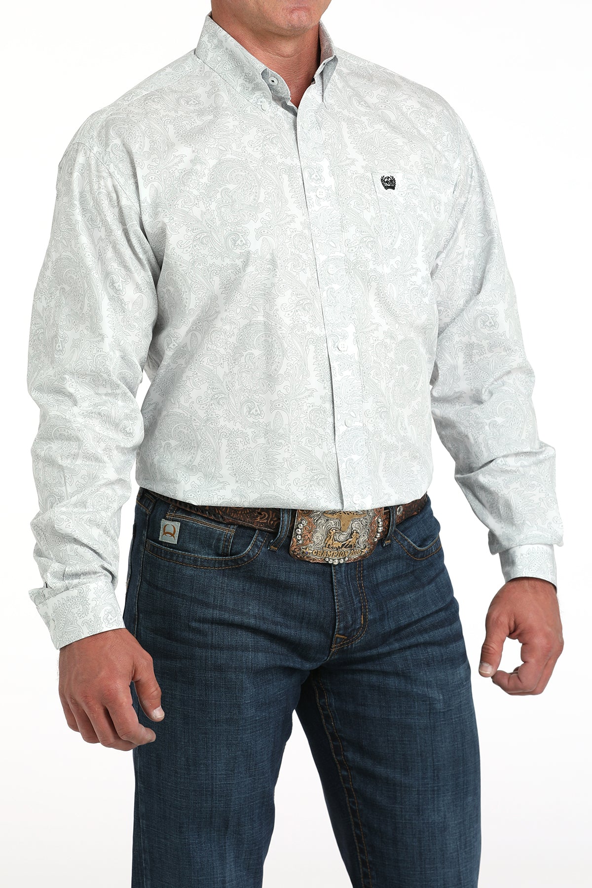 A man wearing a light patterned button-up shirt, dark jeans, and a large belt buckle stands against a white background