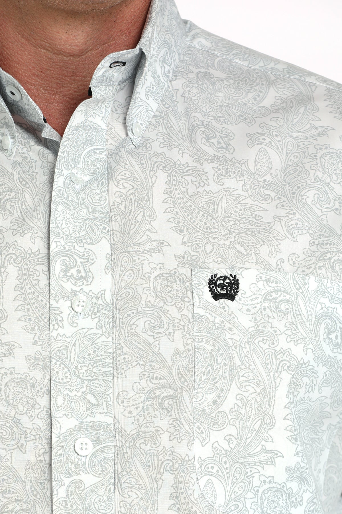Close-up of a man wearing a light patterned button-up shirt with a small black emblem on the chest, displayed against a white background
