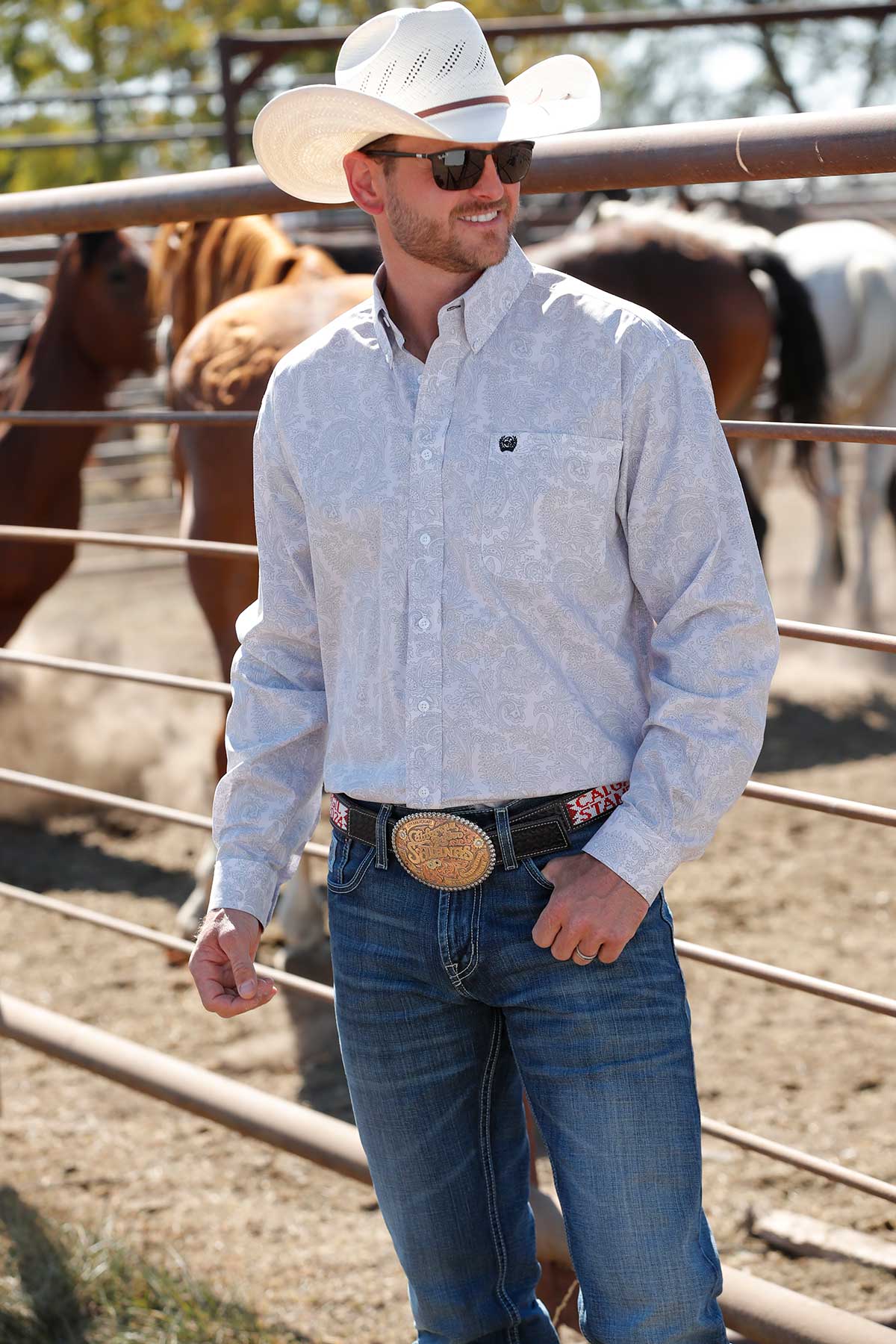 A man wearing a white cowboy hat, patterned long-sleeve shirt, jeans, and a large belt buckle stands by a metal fence with horses in the background