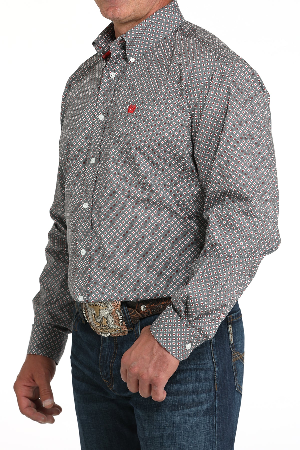 A man wearing a gray patterned button-up shirt, dark jeans, and a large belt buckle stands against a white background