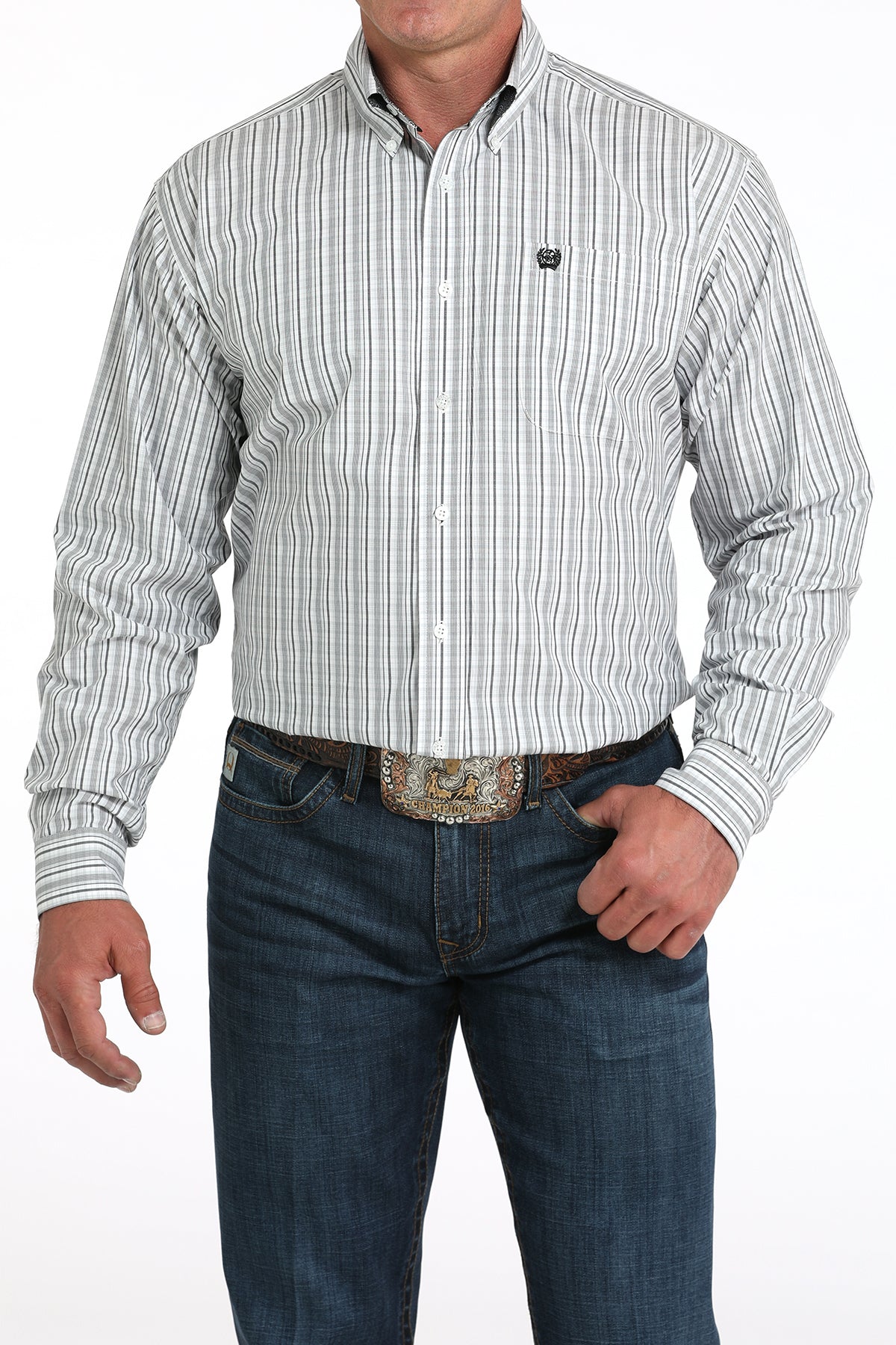 A man wearing a light striped button-up shirt, dark jeans, and a large belt buckle stands against a white background