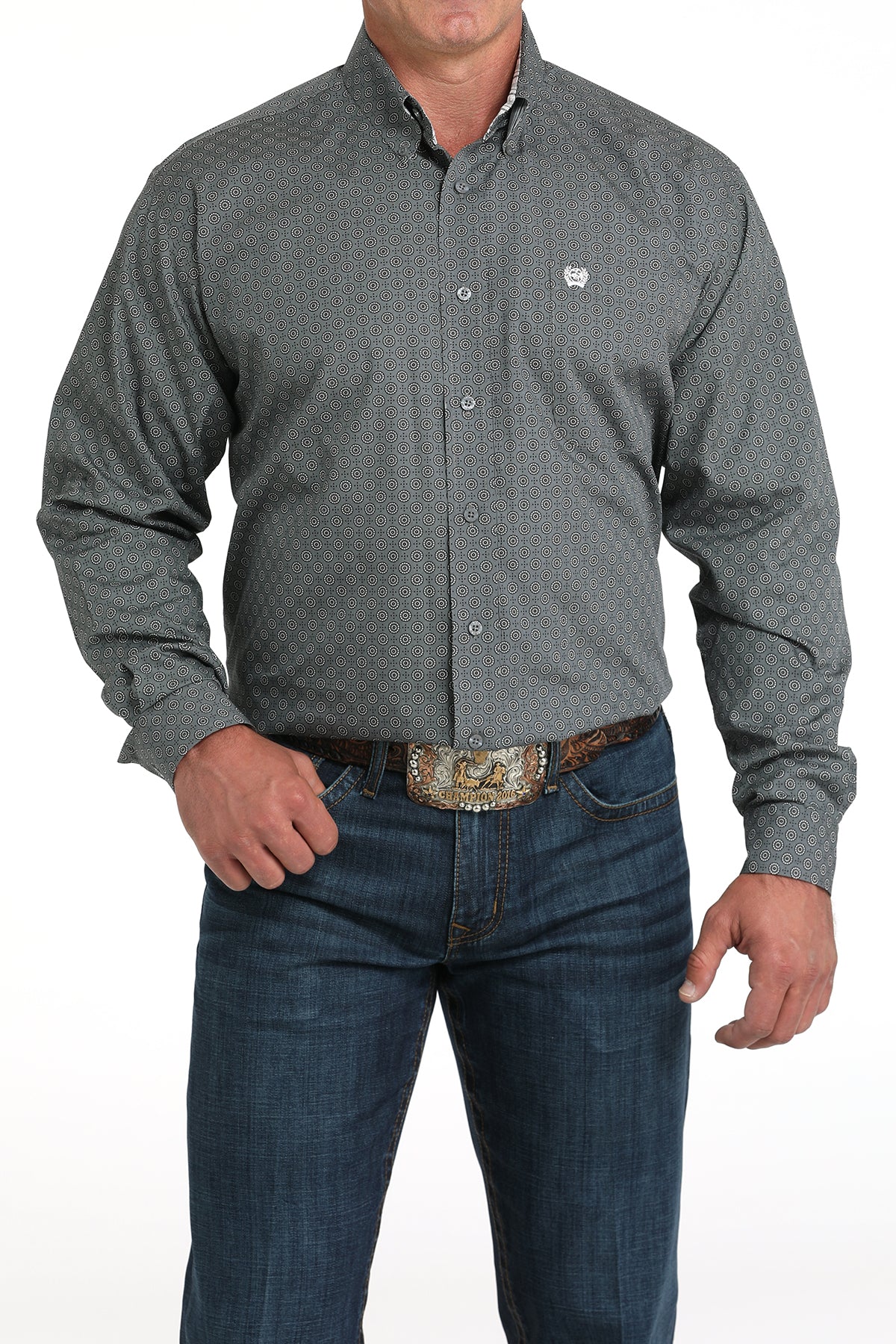 A man wearing a gray patterned button-up shirt, dark jeans, and a large belt buckle stands against a white background