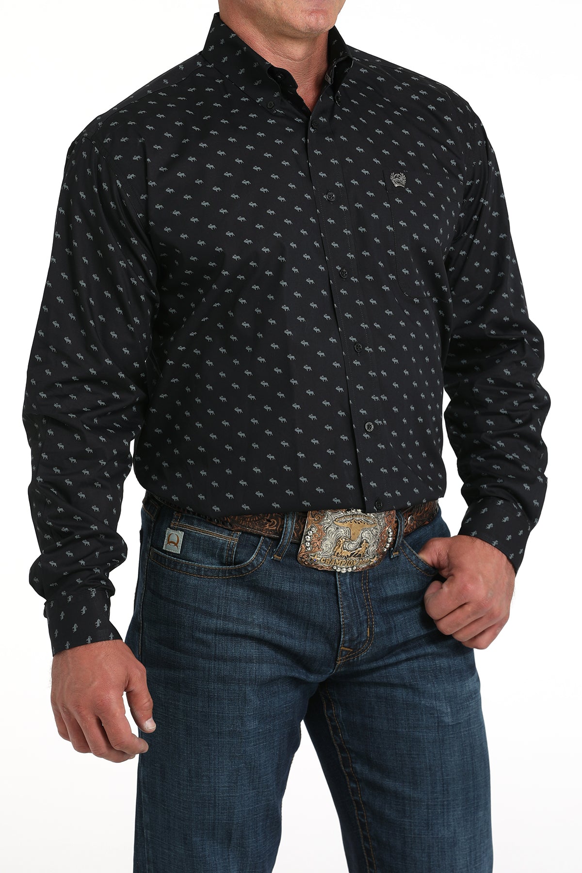 A man wearing a black patterned button-up shirt, dark jeans, and a large belt buckle, standing against a white background