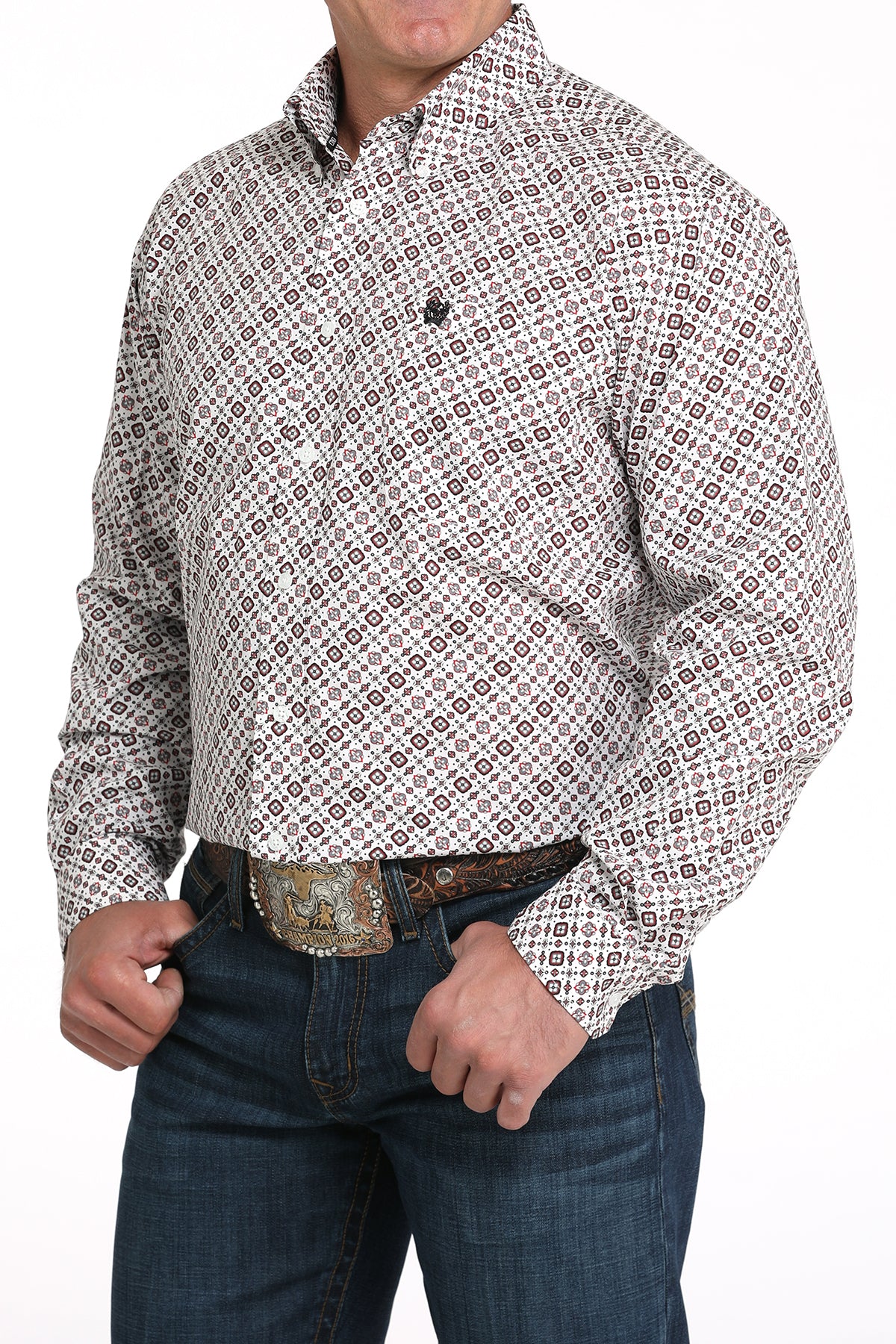 A man wearing a light patterned button-up shirt, dark jeans, and a large belt buckle, standing against a white background
