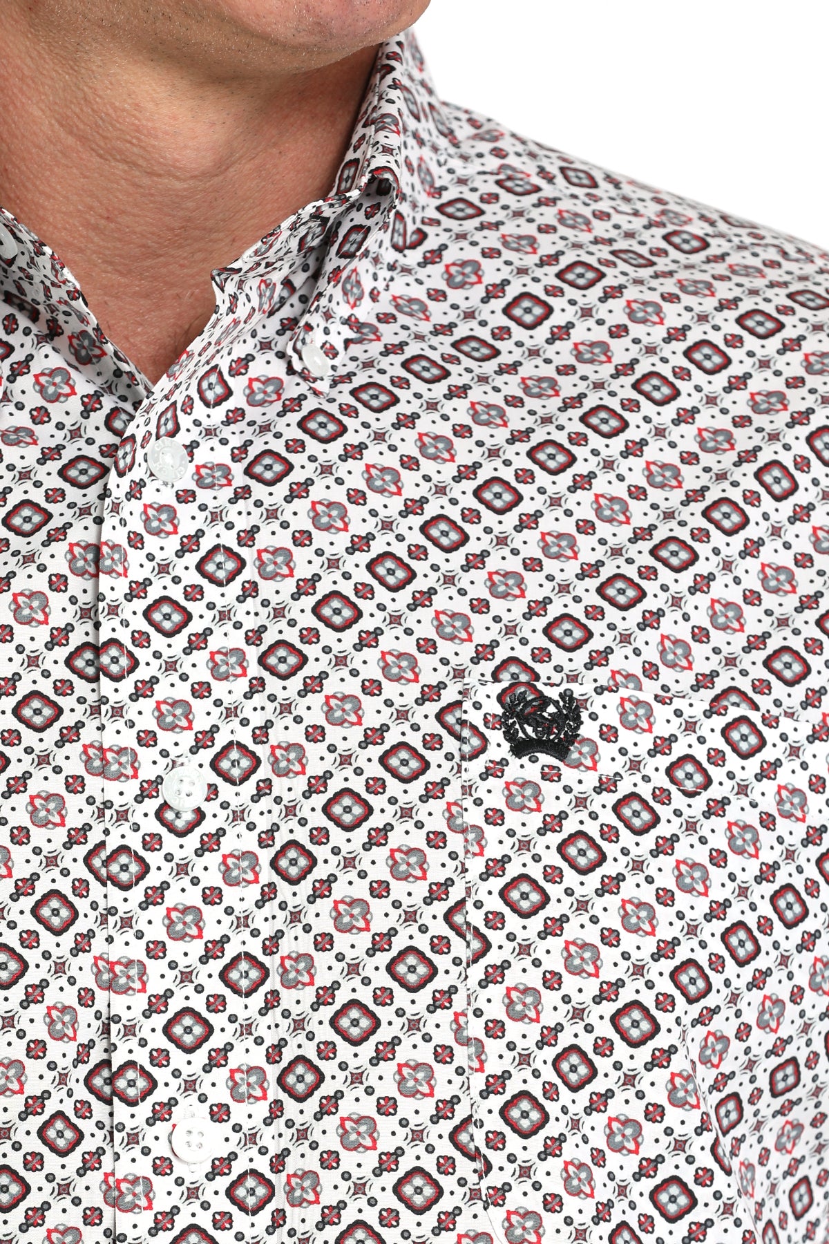 Close-up of a man wearing a light patterned button-up shirt with a small black emblem on the chest, displayed against a white background