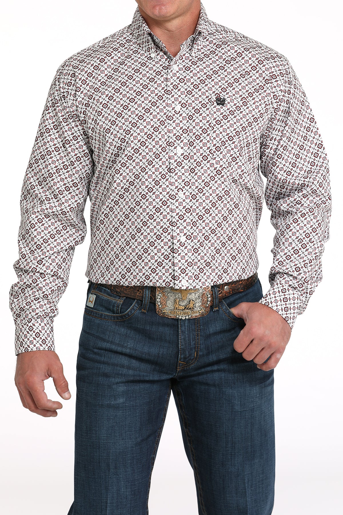 A man wearing a light patterned button-up shirt, dark jeans, and a large belt buckle, standing against a white background
