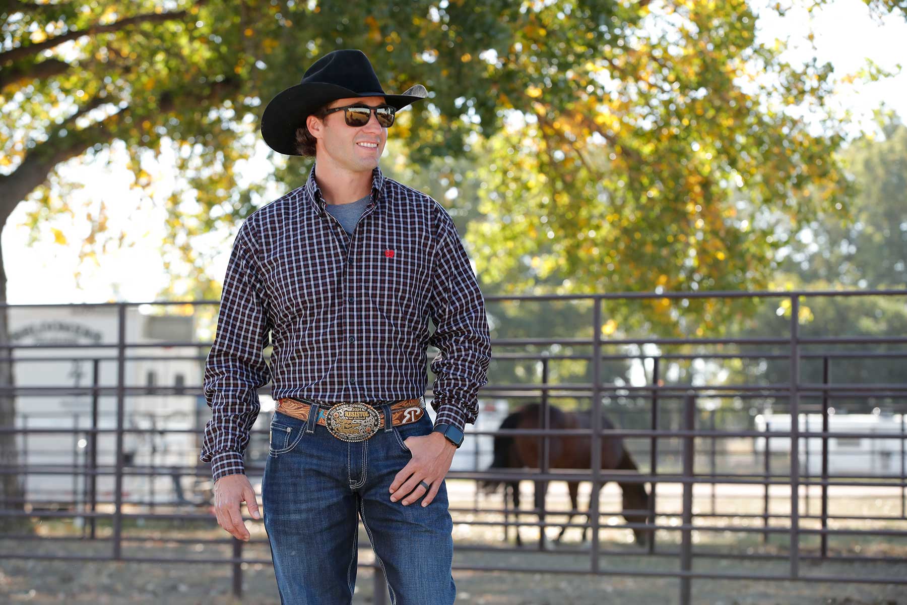A man wearing a black cowboy hat, black patterned long-sleeve shirt, and jeans stands outside near a fence with a trailer and horses in the background