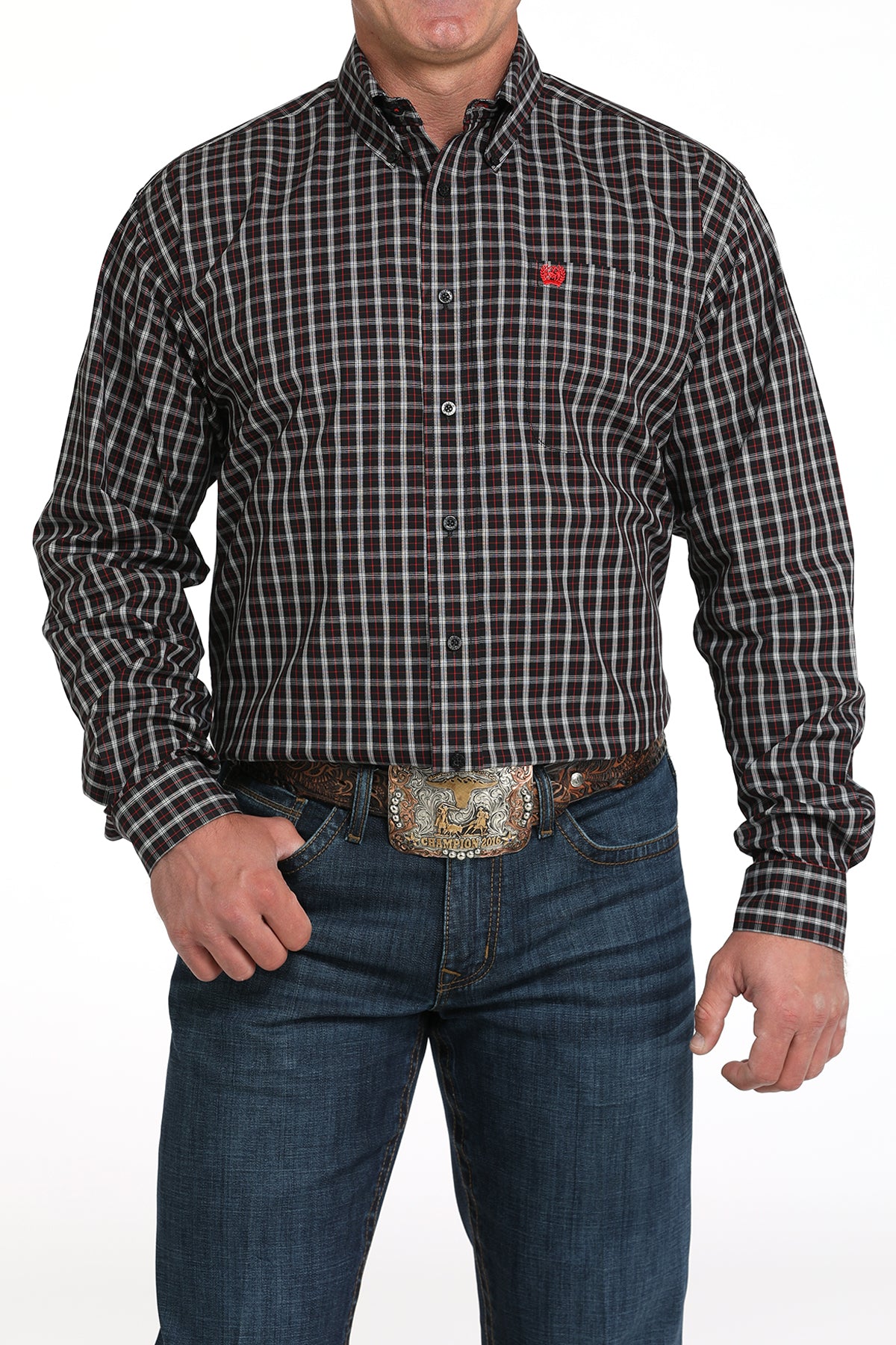 A man wearing a black patterned button-up shirt, dark jeans, and a large belt buckle, standing against a white background