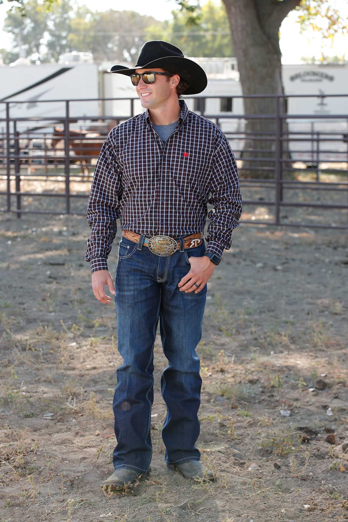 A man wearing a black cowboy hat, black patterned long-sleeve shirt, and jeans stands outside near a fence with a trailer and horses in the background