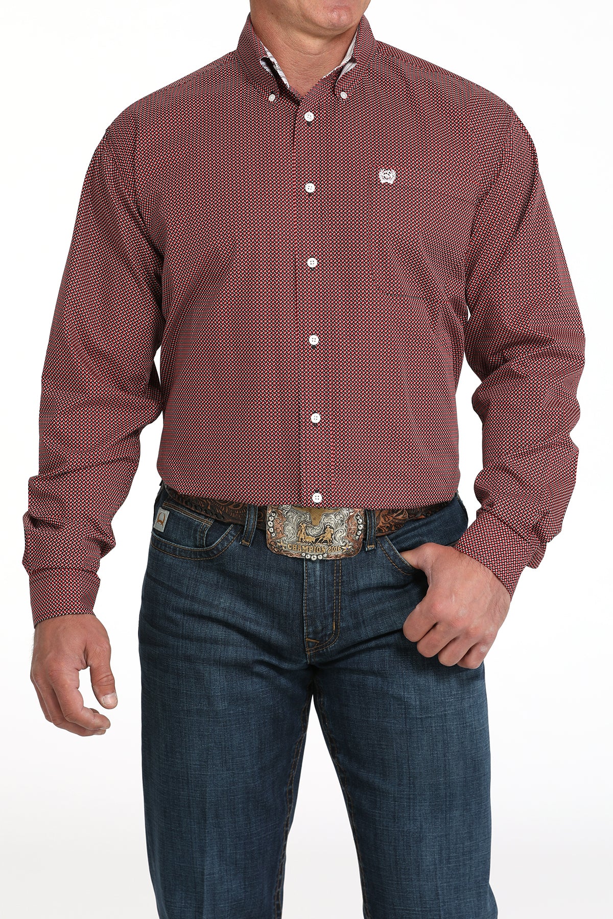A man wearing a maroon patterned button-up shirt, dark jeans, and a large belt buckle, standing against a white background