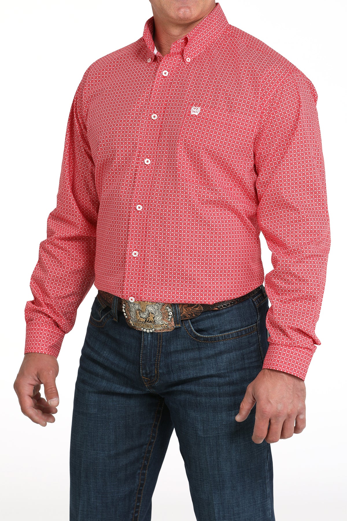 A man wearing a red patterned button-up shirt, dark jeans, and a large belt buckle, standing against a white background