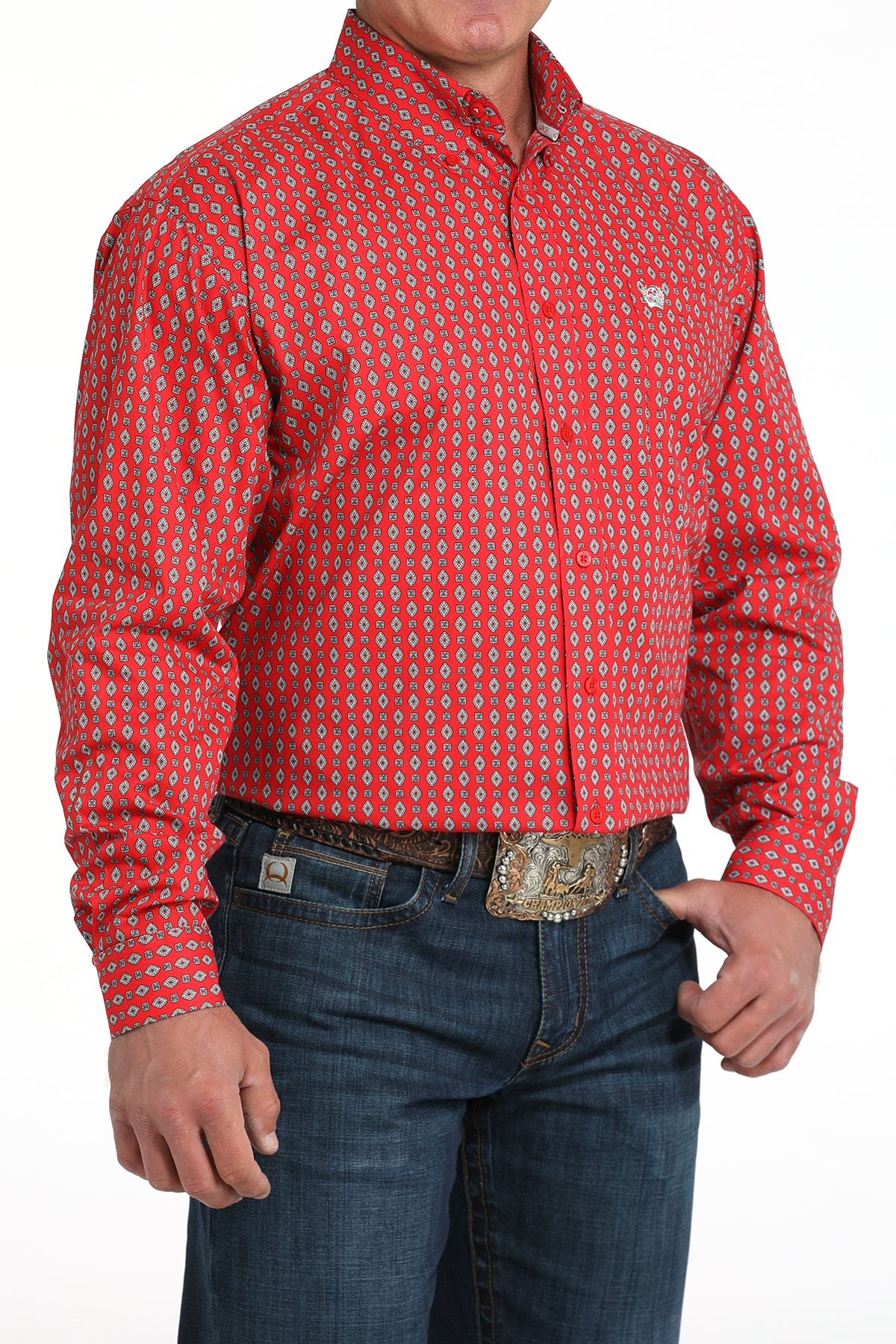 A man wearing a red patterned button-up shirt, dark jeans, and a large belt buckle, standing against a white background