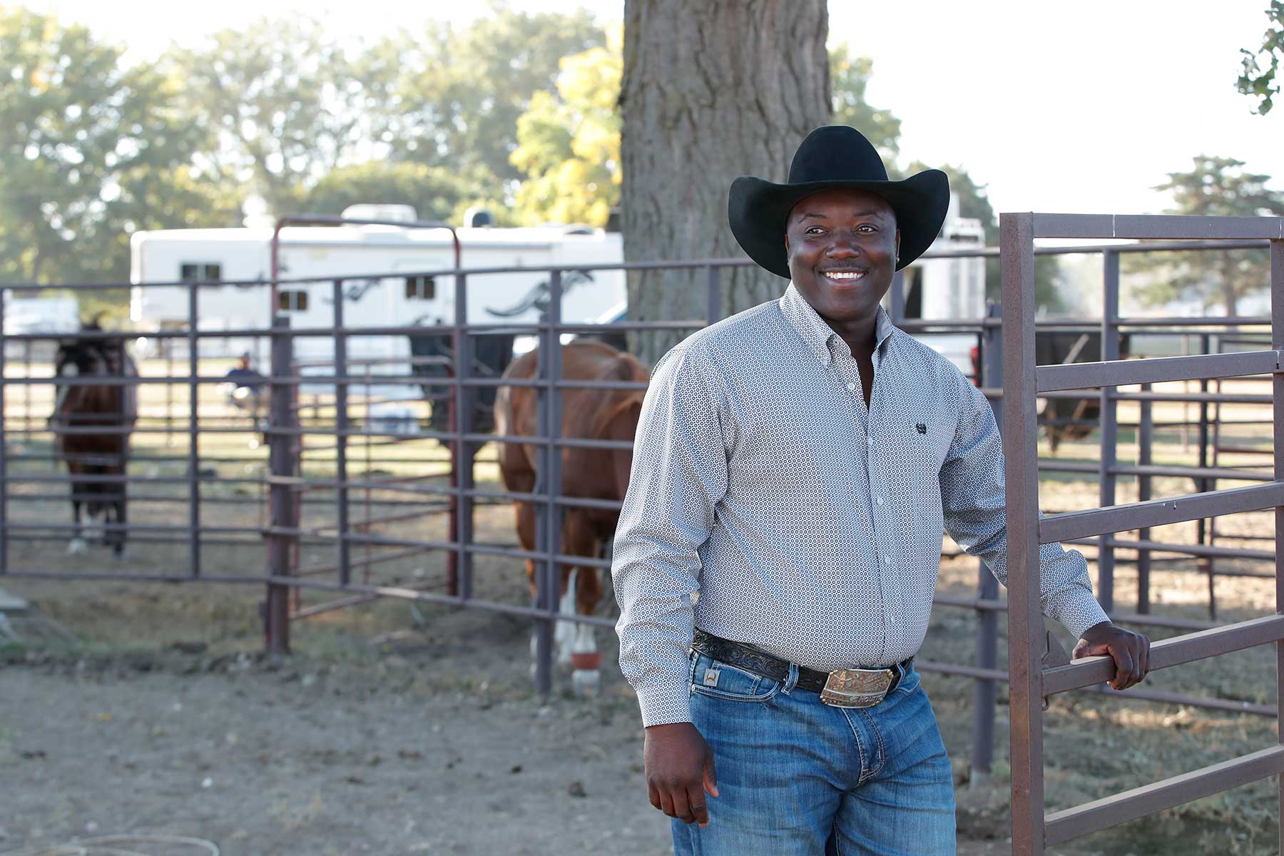 Man standing outside in front of a corral with horses, wearing a black cowboy hat, long sleeve button-down shirt and jeans with a belt buckle 