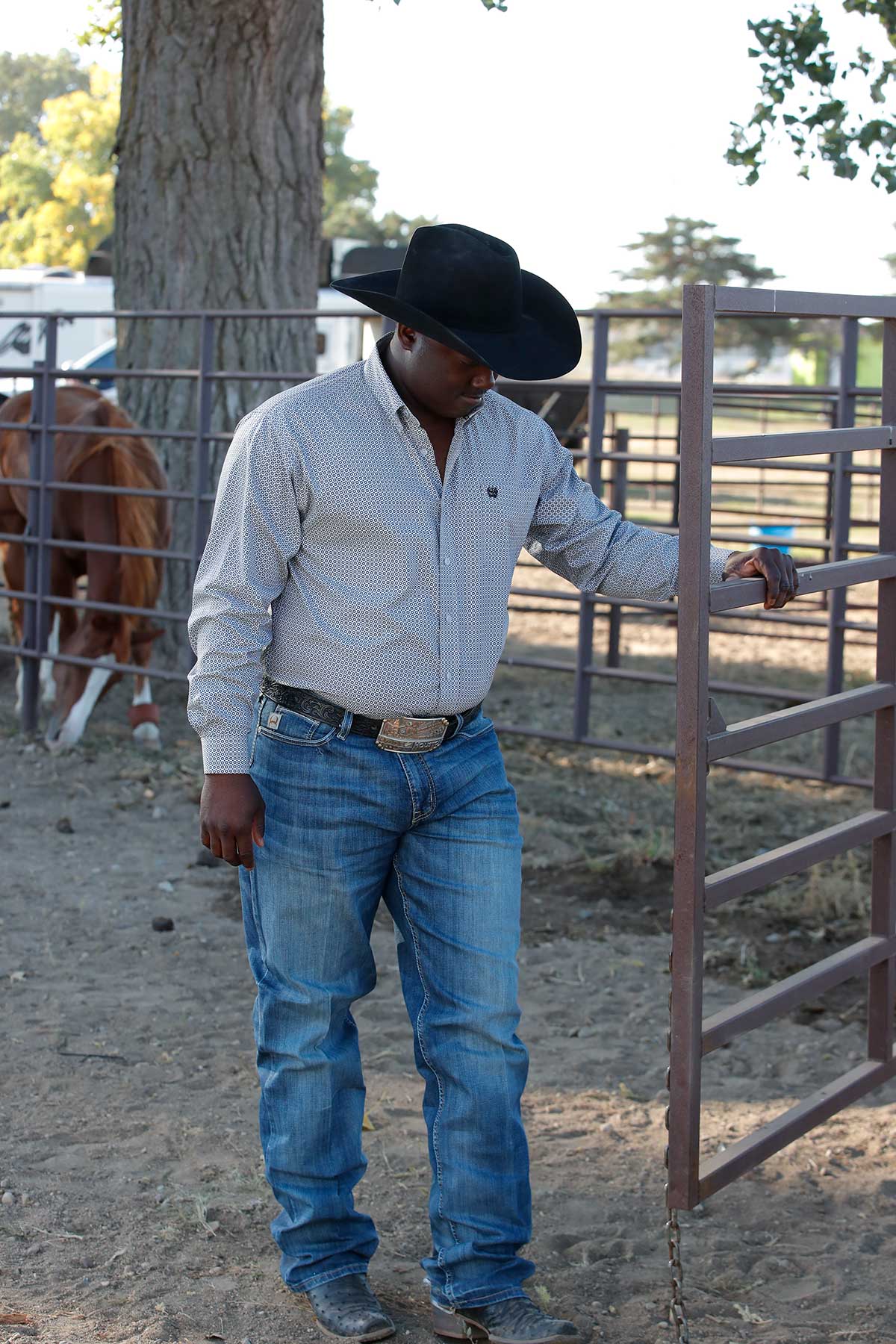 Man standing outside in front of a corral with horses, wearing a black cowboy hat, long sleeve button-down shirt and jeans with a belt buckle 