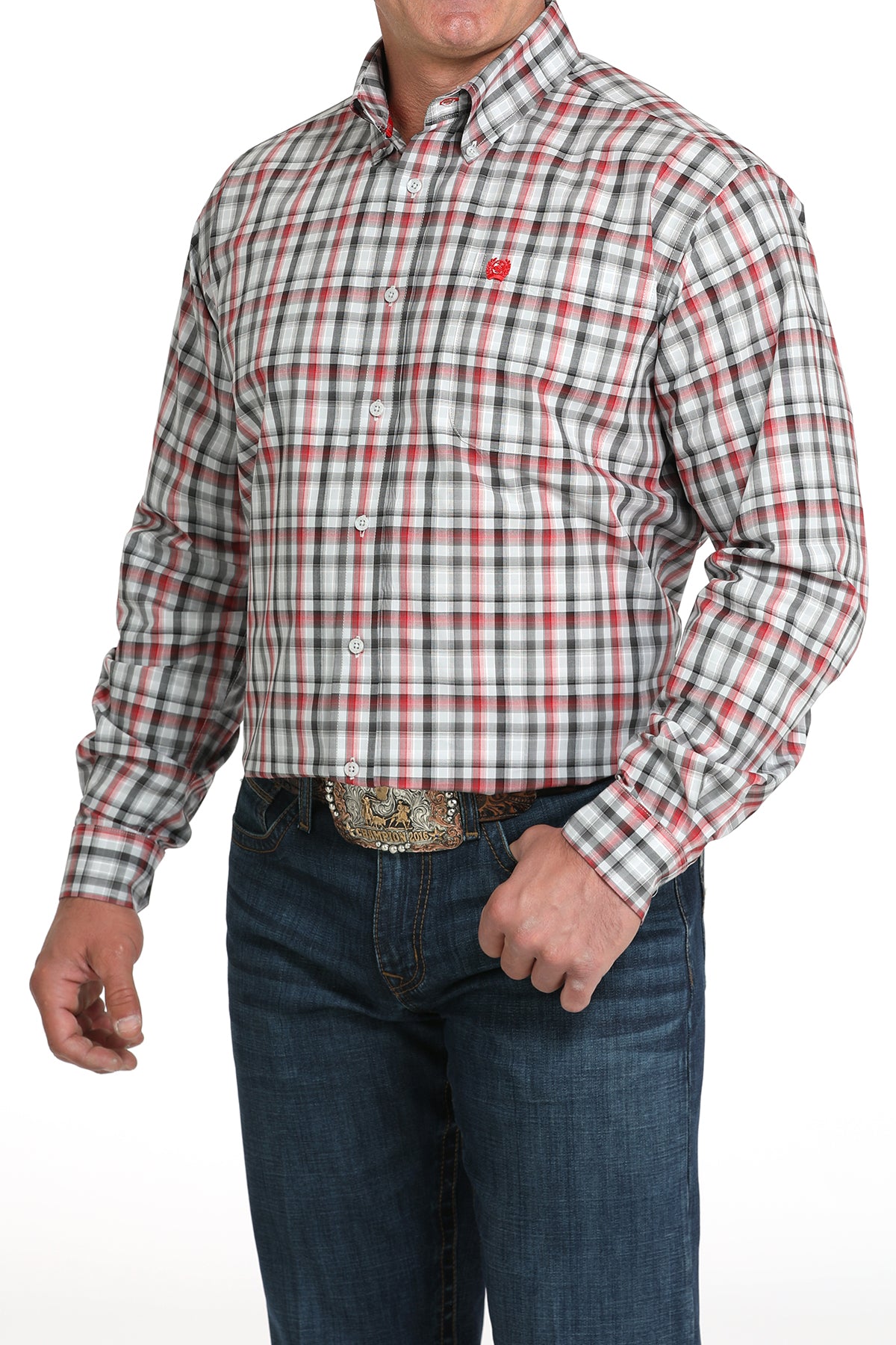 A man wearing a red, white and black plaid button-up shirt, dark jeans, and a large belt buckle, standing against a white background