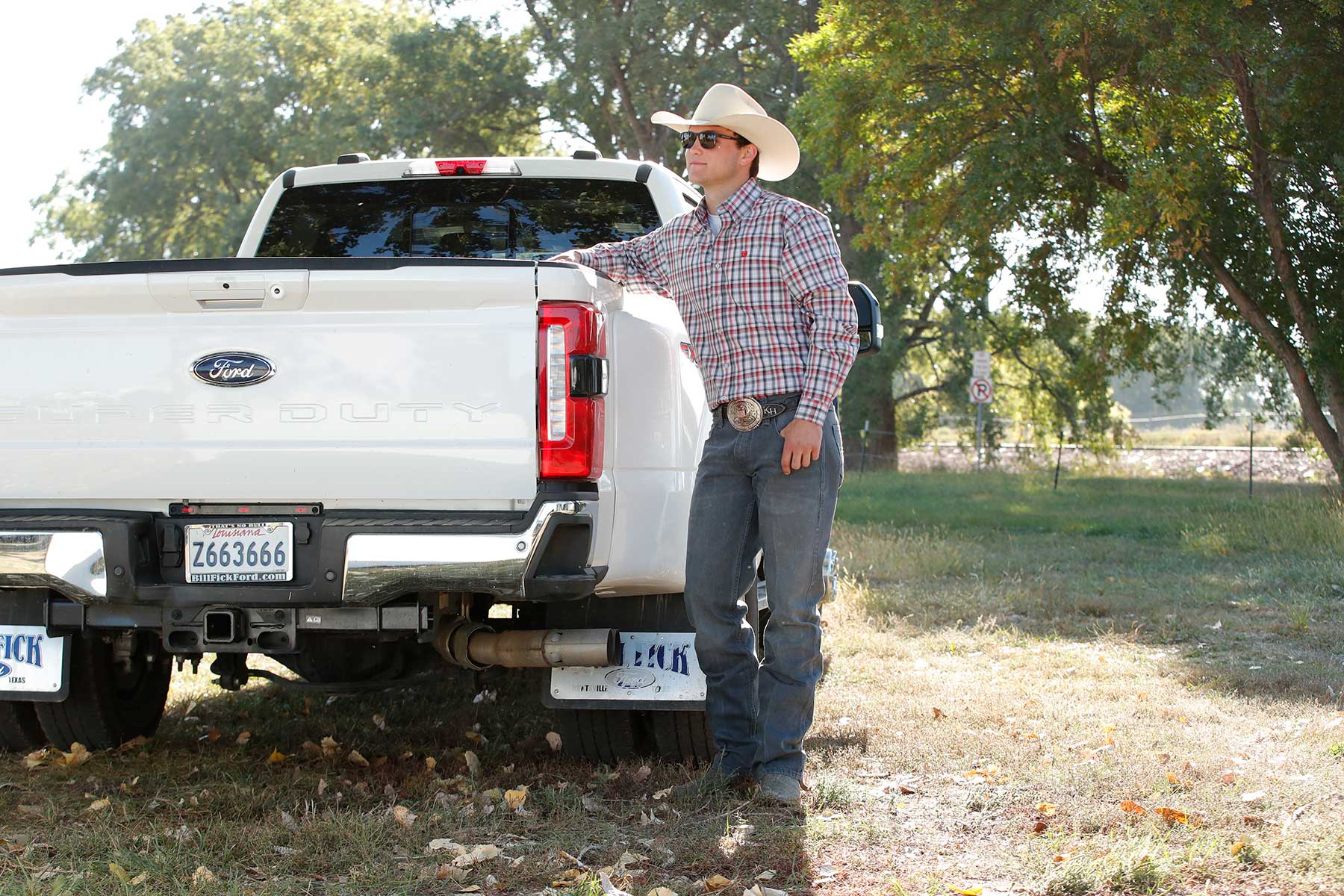 Man standing outside, leaning against a truck wearing a cowboy hat, plaid shirt and jeans