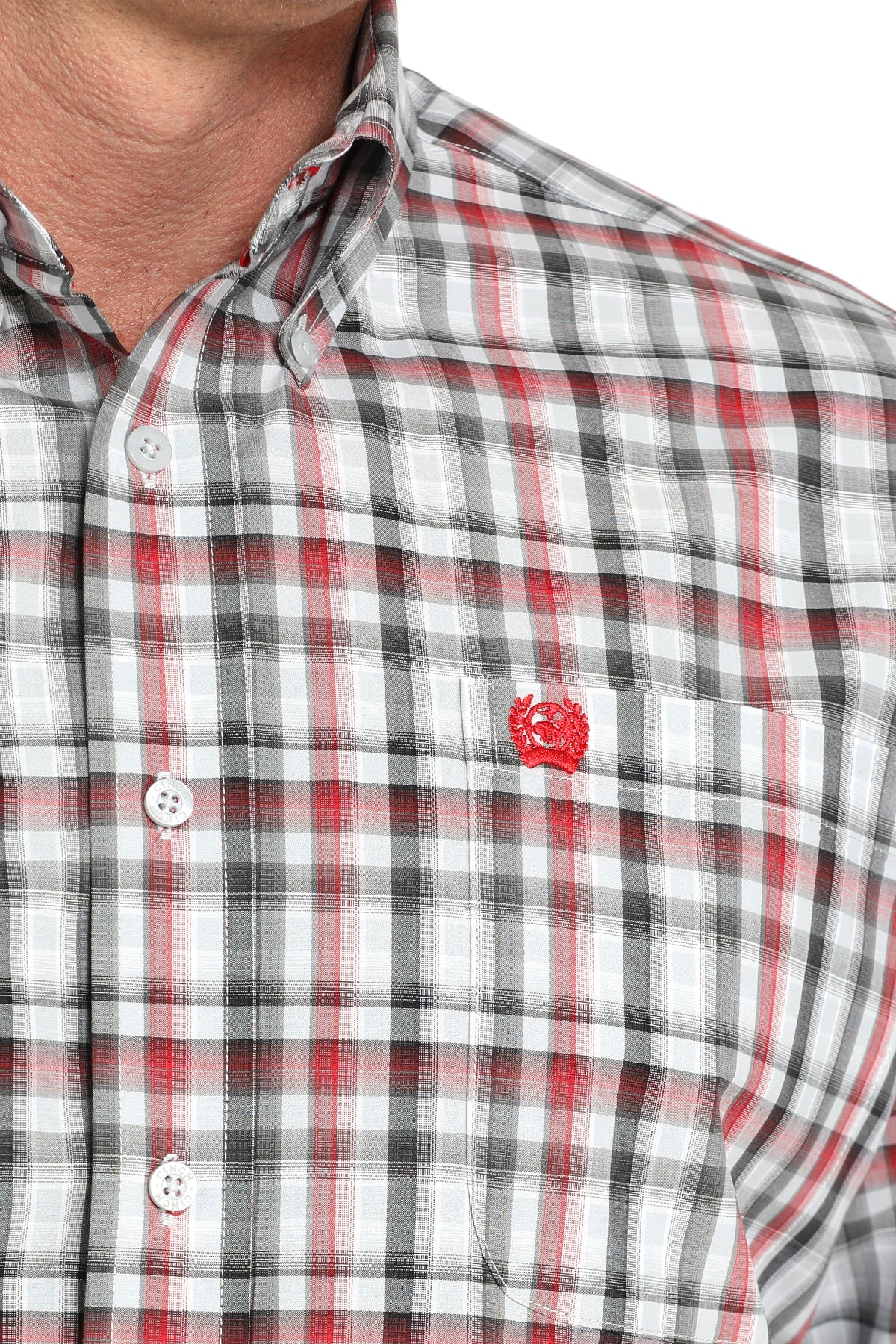 Close-up of a man wearing a red, white and black plaid button-up shirt with a small red emblem on the chest, displayed against a white background