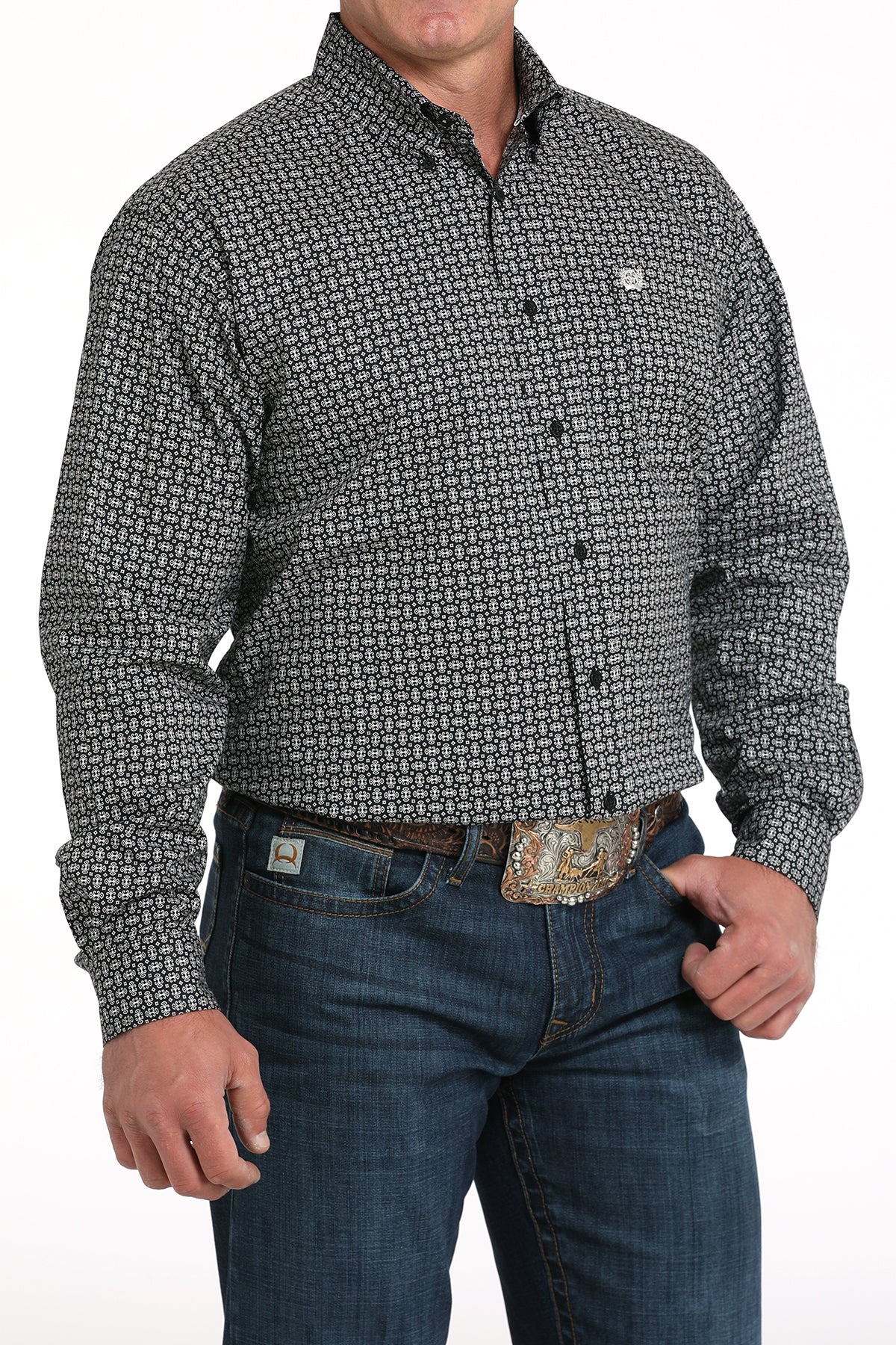 A man wearing a gray patterned button-up shirt, dark jeans, and a large belt buckle, standing against a white background