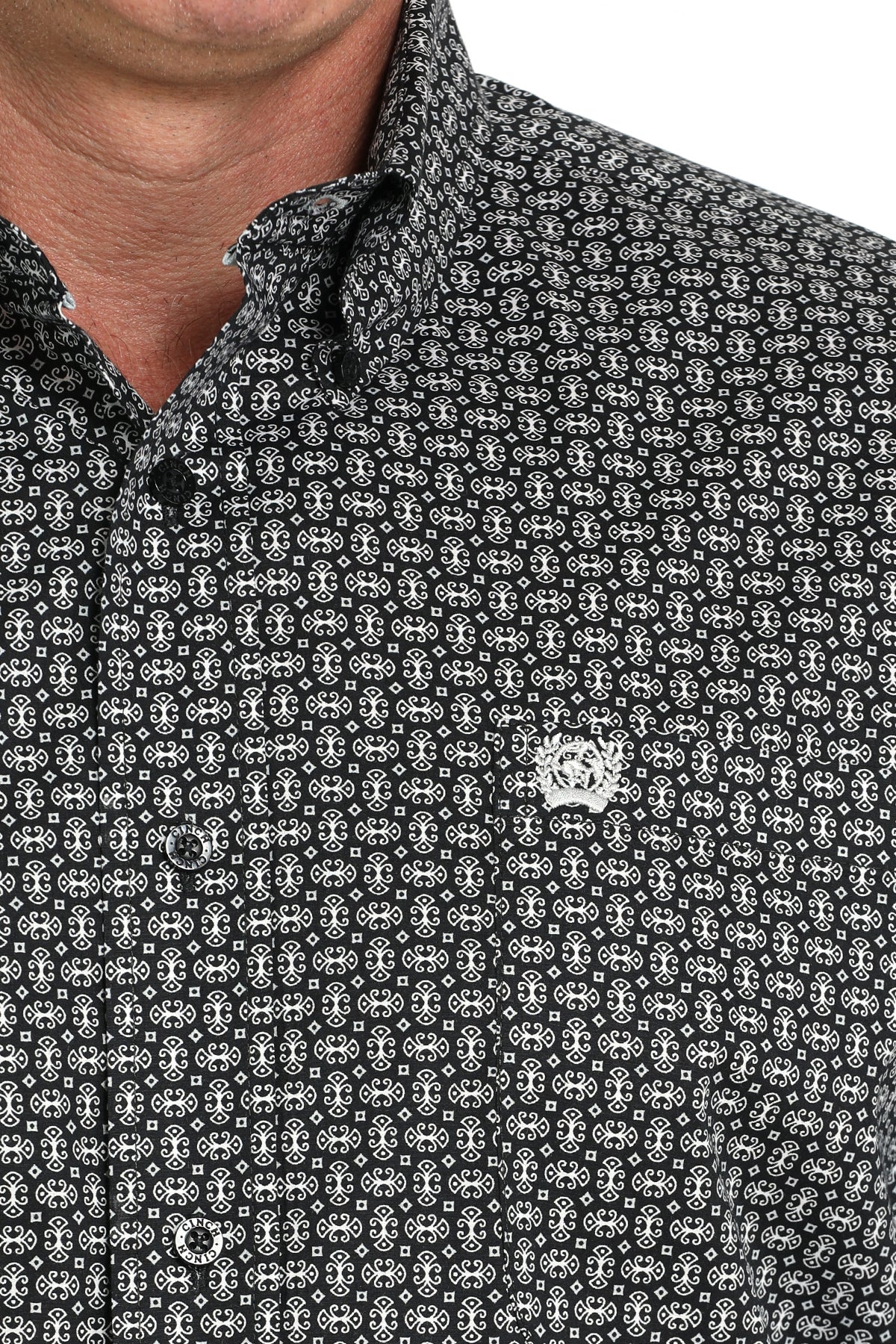 Close-up of a man wearing a gray patterned button-up shirt with a small  emblem on the chest, displayed against a white background