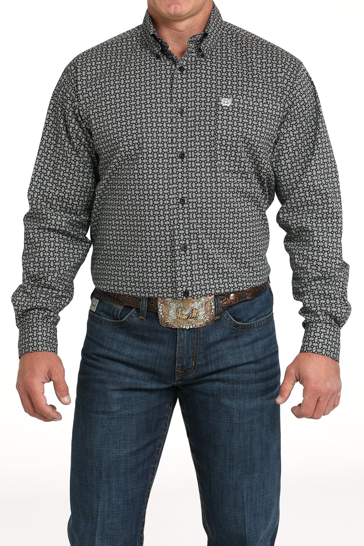 A man wearing a gray patterned button-up shirt, dark jeans, and a large belt buckle, standing against a white background