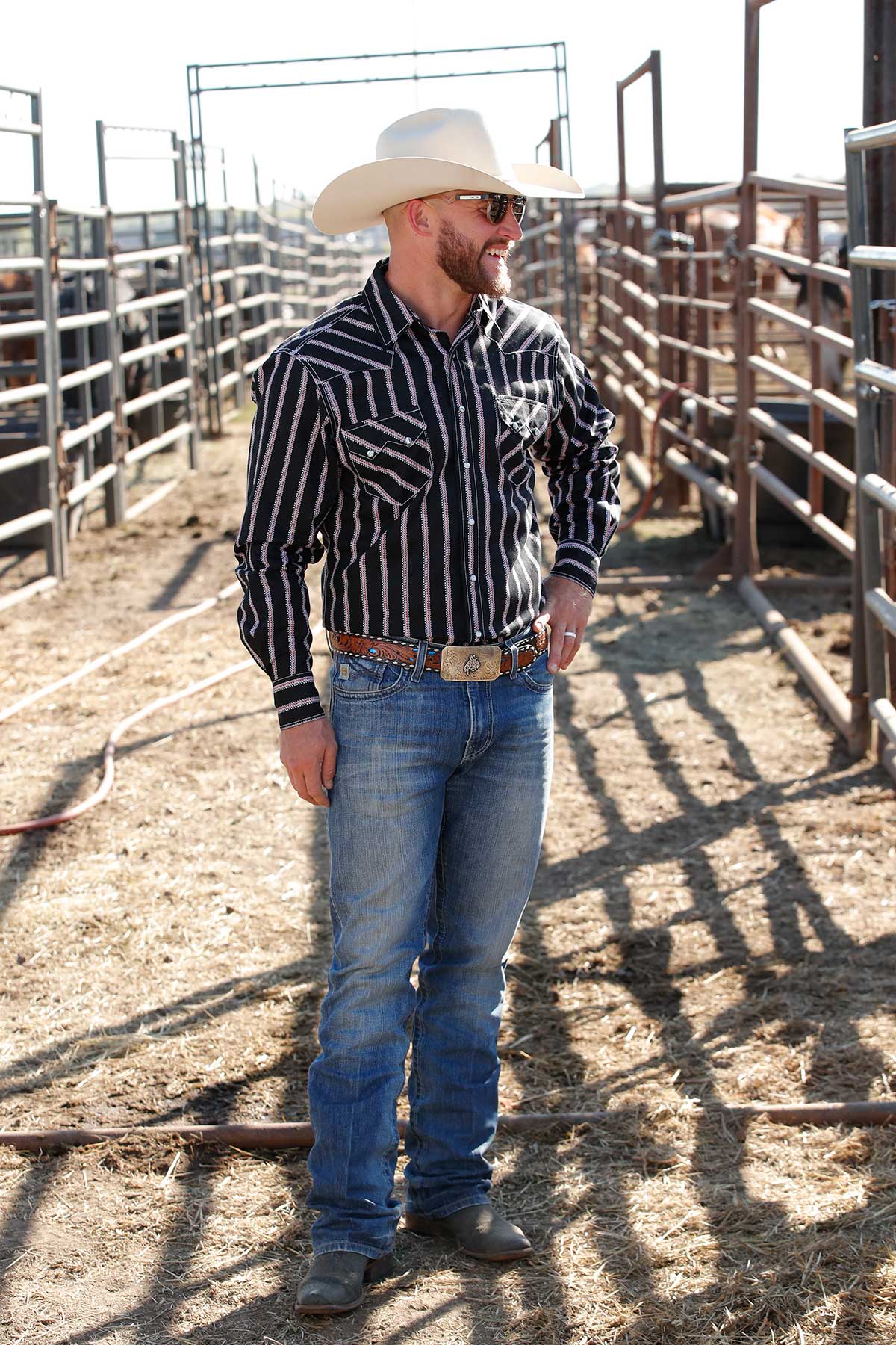 Man wearing a cream cowboy hat, sunglasses, black striped button-down shirt, blue jeans, and a silver belt buckle. He is standing in a cattle pen with metal fencing and looking off to the side