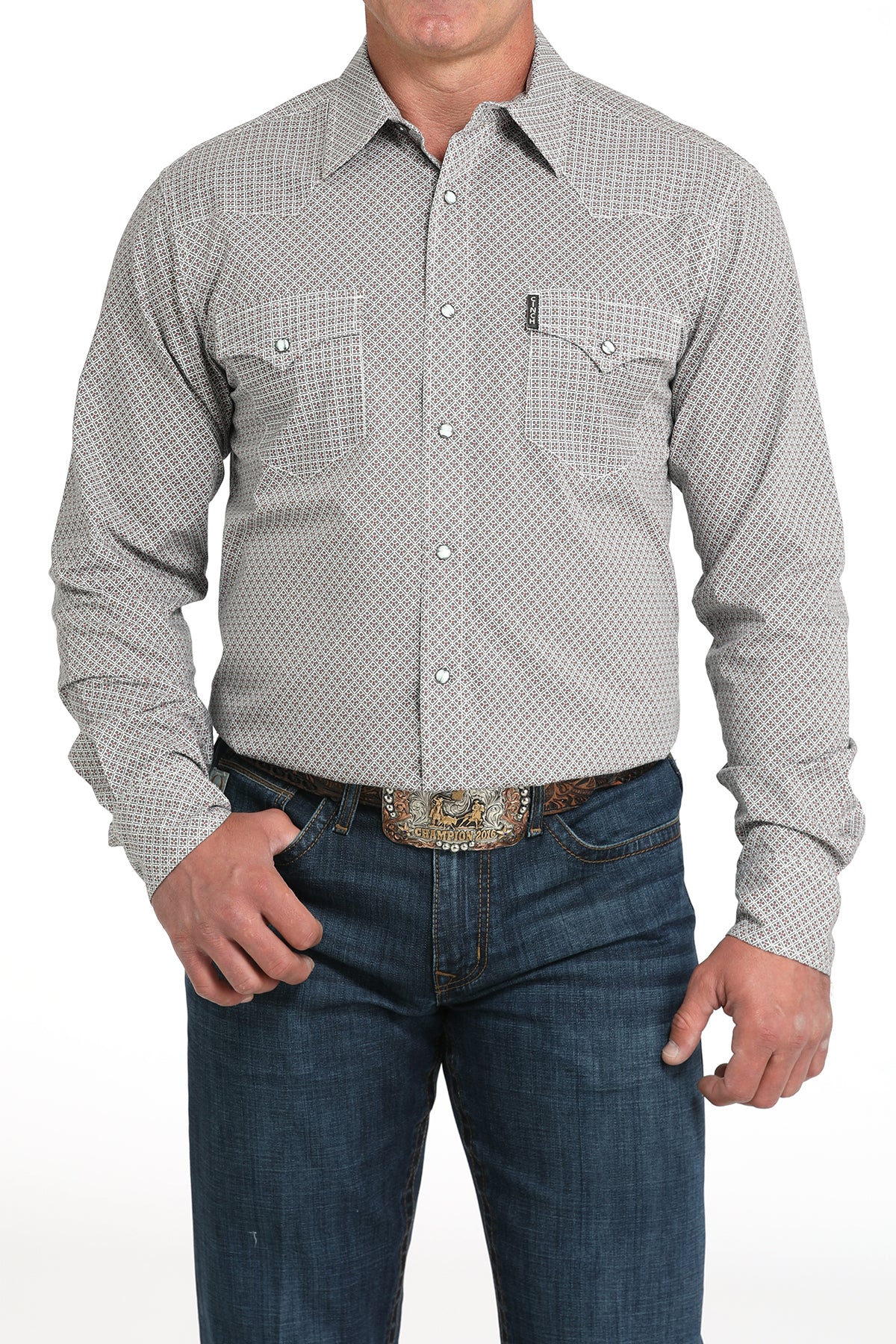 A man wearing a light gray patterned button-up shirt, dark jeans, and a large belt buckle, standing against a white background
