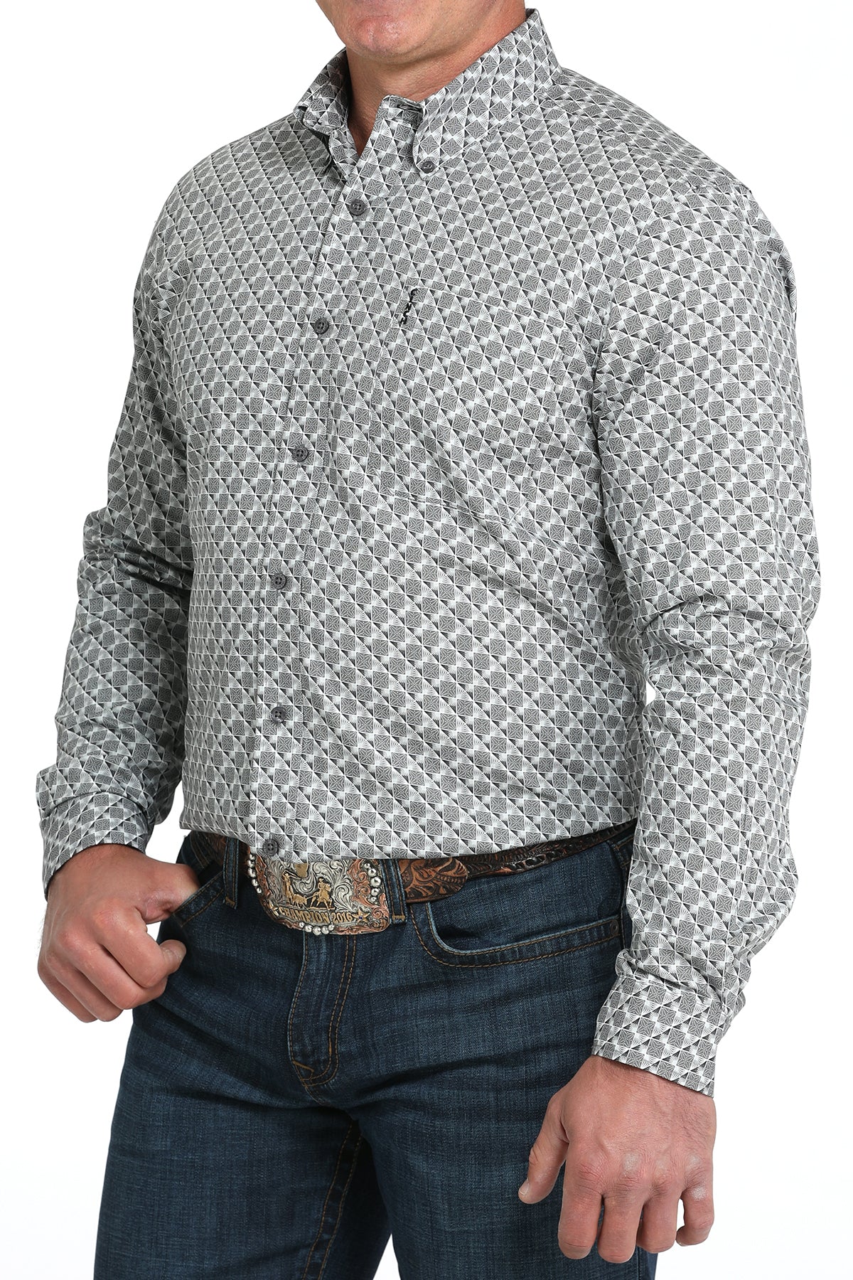 A man wearing a light gray patterned button-up shirt, dark jeans, and a large belt buckle, standing against a white background