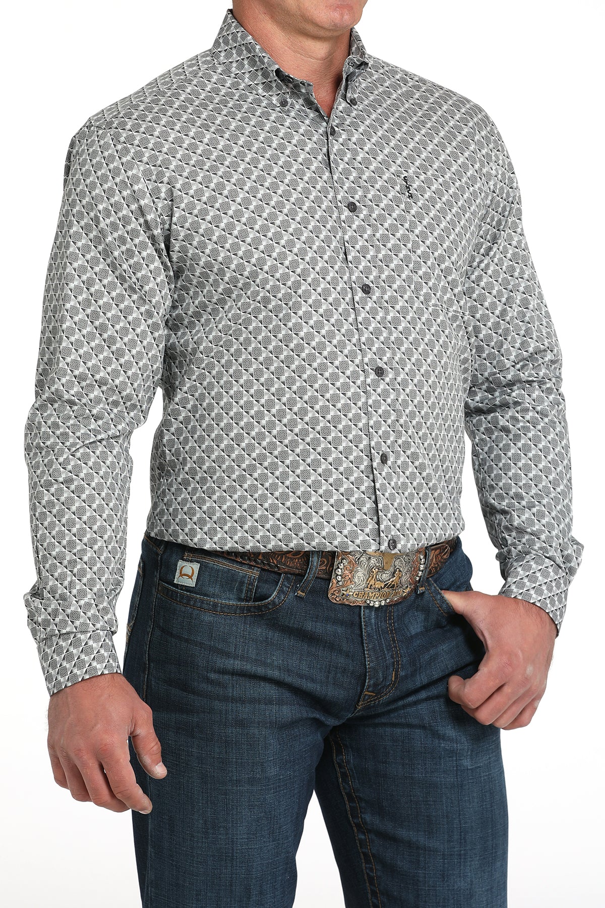 A man wearing a light gray patterned button-up shirt, dark jeans, and a large belt buckle, standing against a white background