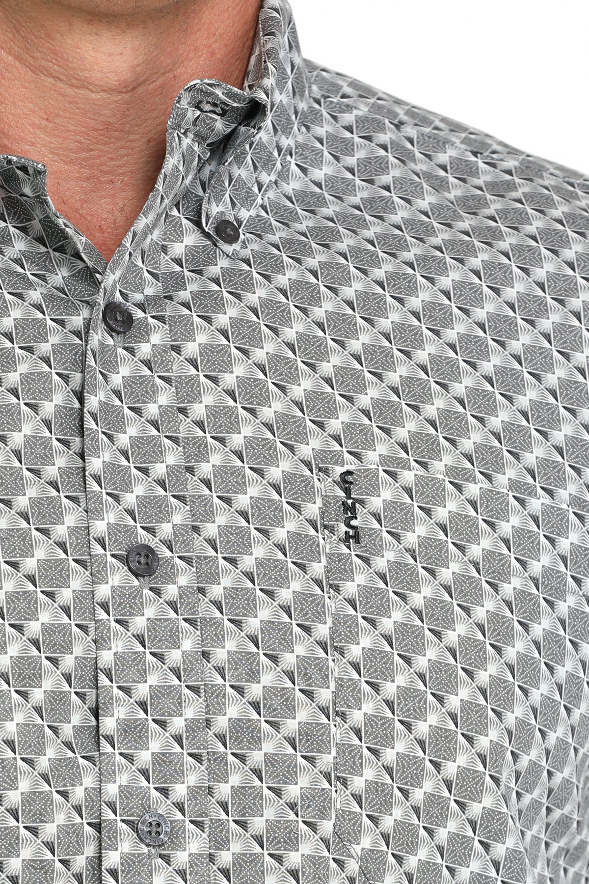 Close-up of a man wearing a gray patterned button-up shirt with a small  emblem on the chest, displayed against a white background
