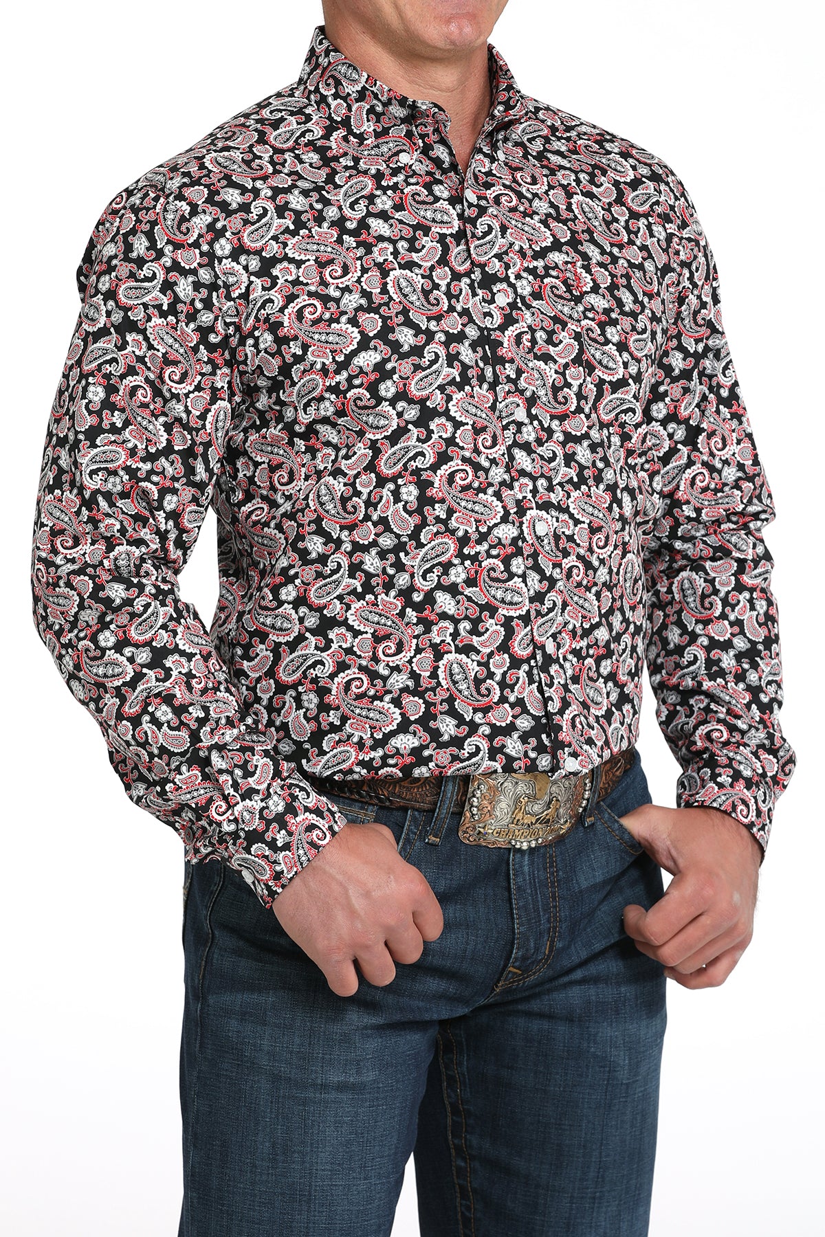 A man wearing a red and black patterned button-up shirt, dark jeans, and a large belt buckle, standing against a white background