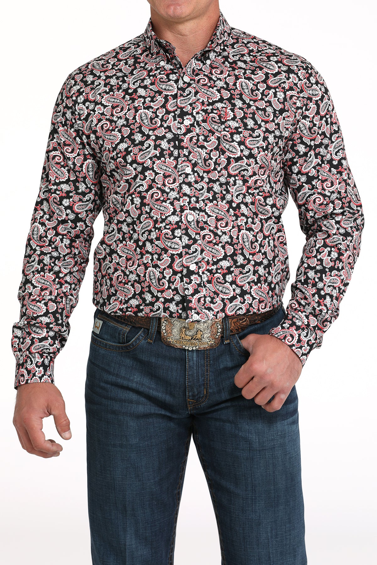 A man wearing a red and black patterned button-up shirt, dark jeans, and a large belt buckle, standing against a white background
