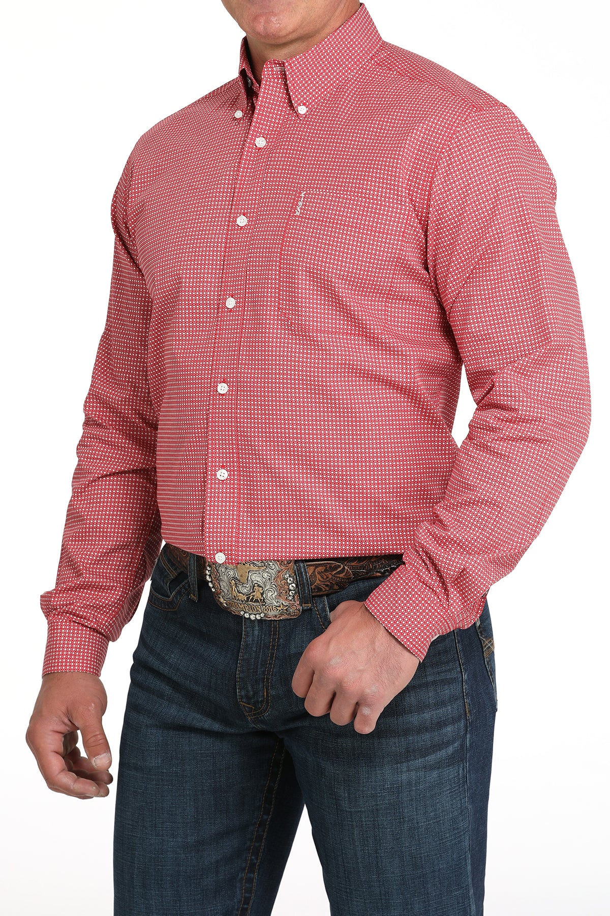 A man wearing a red patterned button-up shirt, dark jeans, and a large belt buckle, standing against a white background