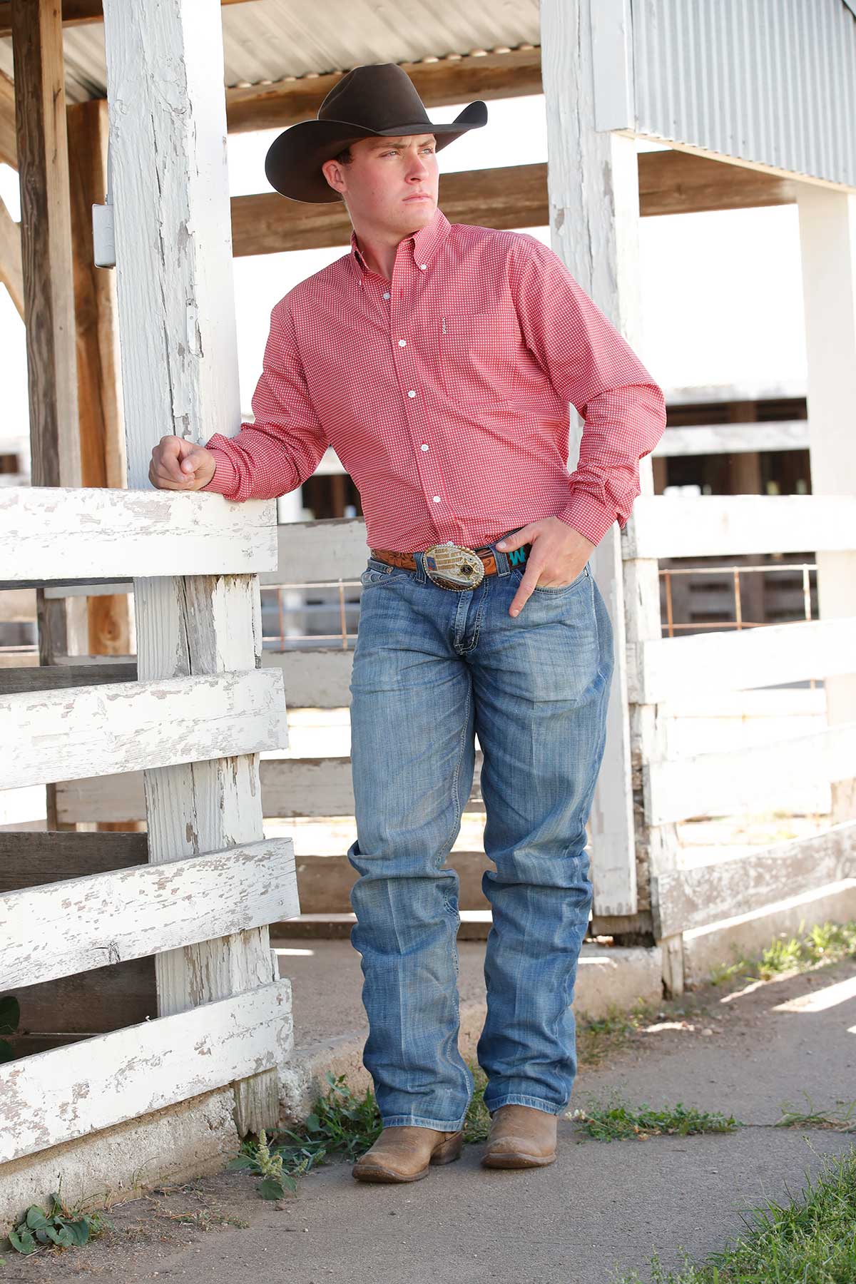 Man wearing a black cowboy hat, red button-up shirt with a small check pattern, blue jeans, and a large belt buckle. He is standing near a white wooden fence in a rustic barn setting, looking off to the side