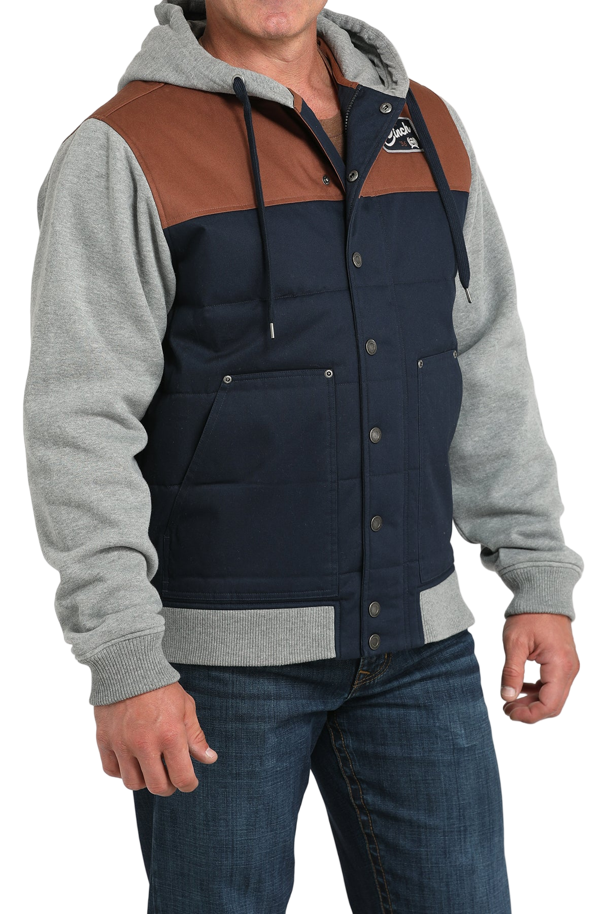 Man wearing a navy and brown snap-front jacket with light gray sleeves and a hood, paired with blue jeans