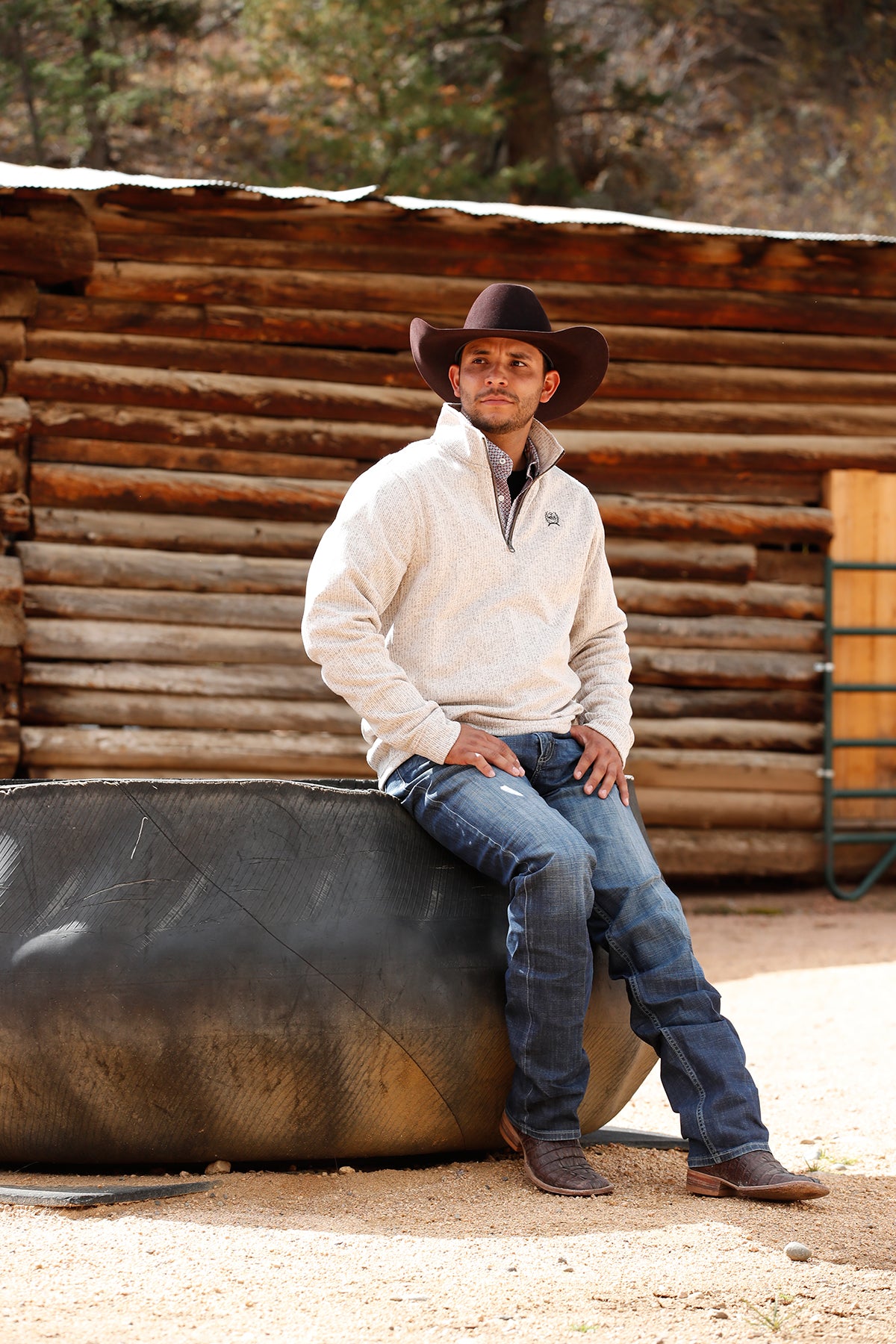 Man wearing a khaki 1/4 zip sweater with jeans and a cowboy hat, leaning on a metal structure outside 