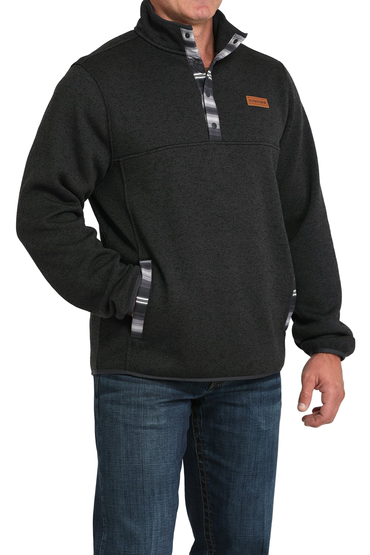 Man wearing a dark textured pullover with patterned trim on the placket and pockets, paired with blue jeans